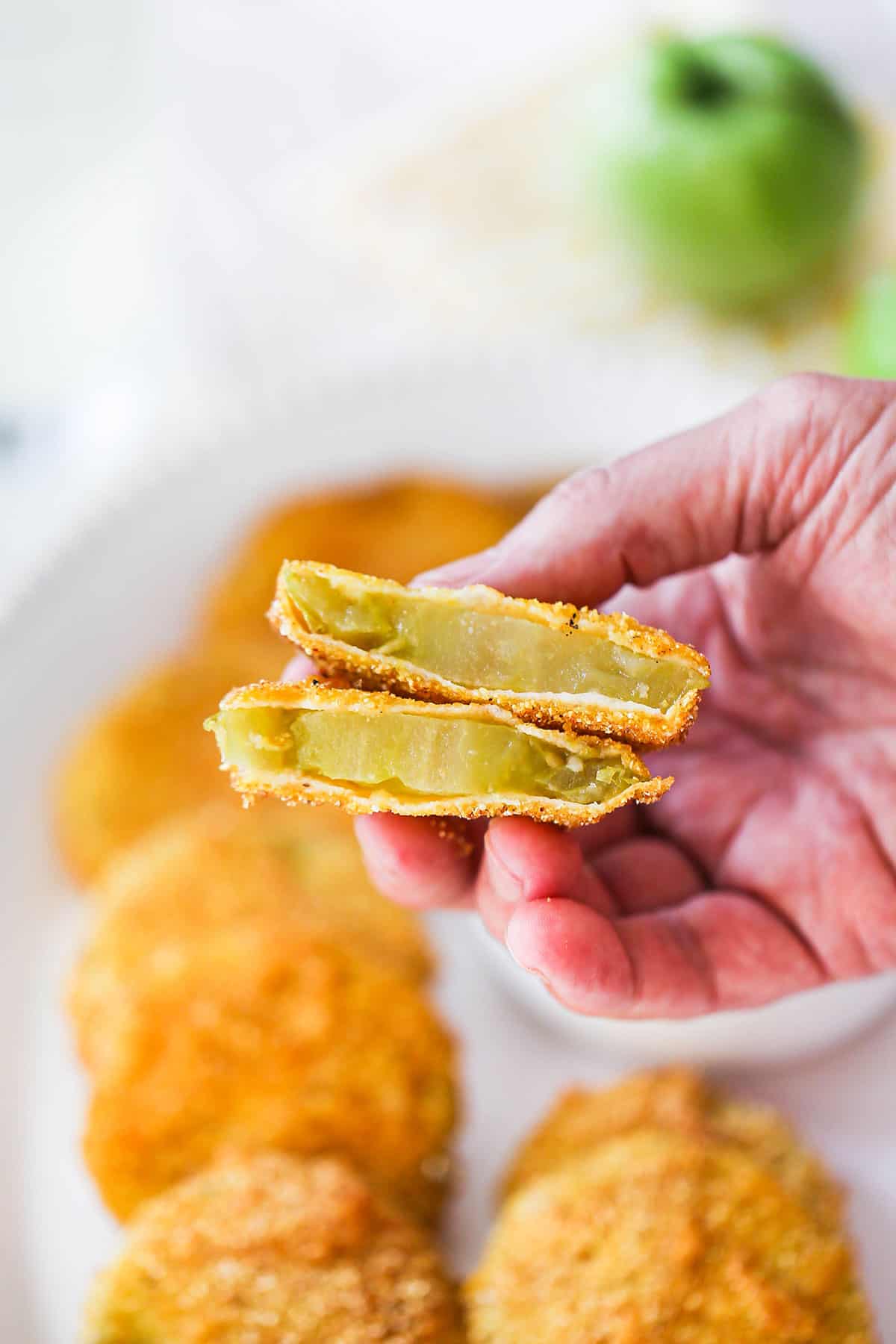 A person holding up two halves of a fried green tomato so the green interior is visible as well as a platter of the fried tomatoes nearby.