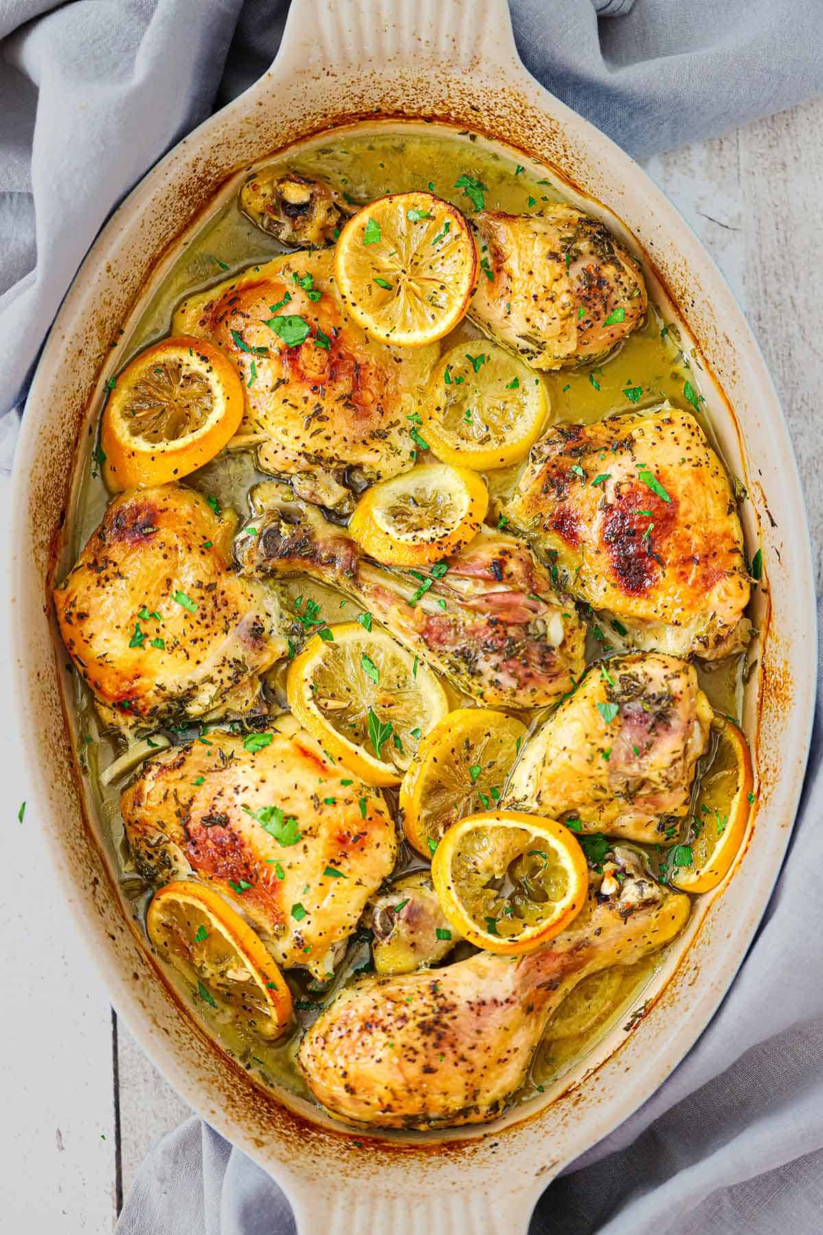 An oval baking dish filled with lemon roast chicken with slices of roasted lemons and lightly browned chicken pieces garnished with chopped Italian parsley.