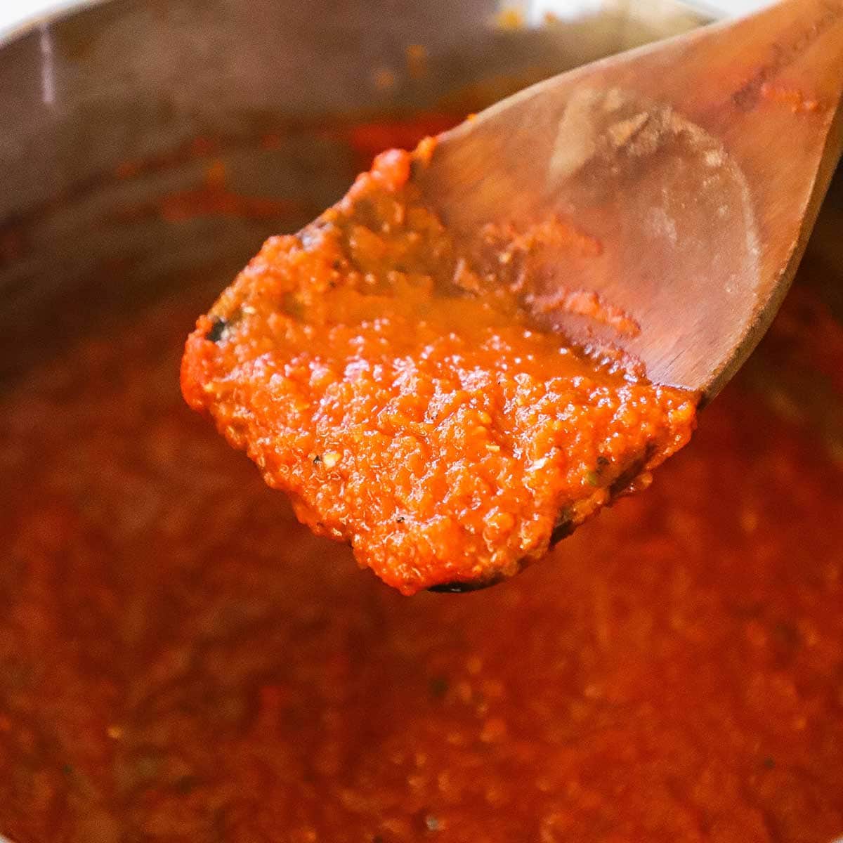 A large wooden spoon being lifted up out of a saucepan filled with homemade ranchero sauce.