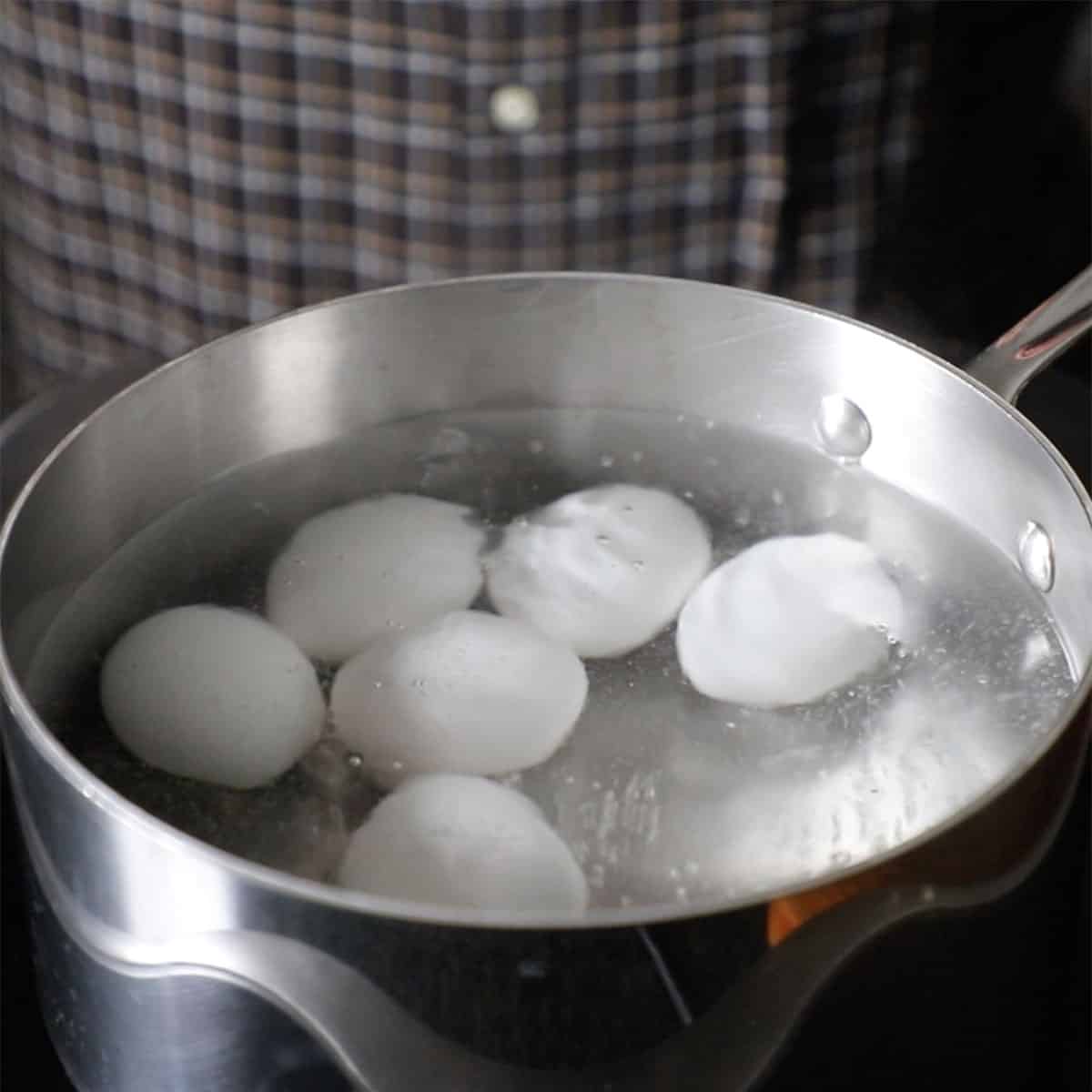 A person standing next to a saucepan filled with simmering water with whole white eggs in the pan.