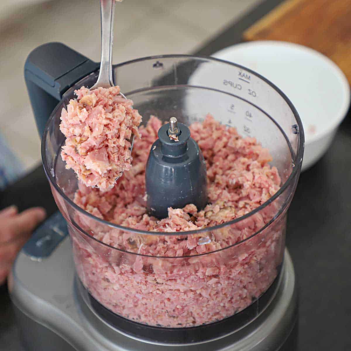 A person holding up chopped ham from the bowl of a food processor that has just finely chopped cooked ham.