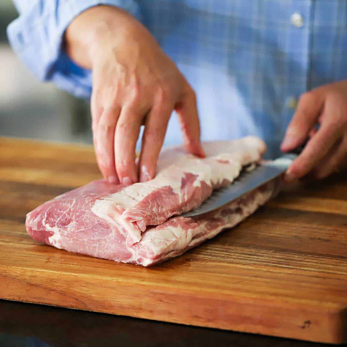 A person using a chef's knife to slice into the top third of an uncooked pork loin roast on a cutting board.