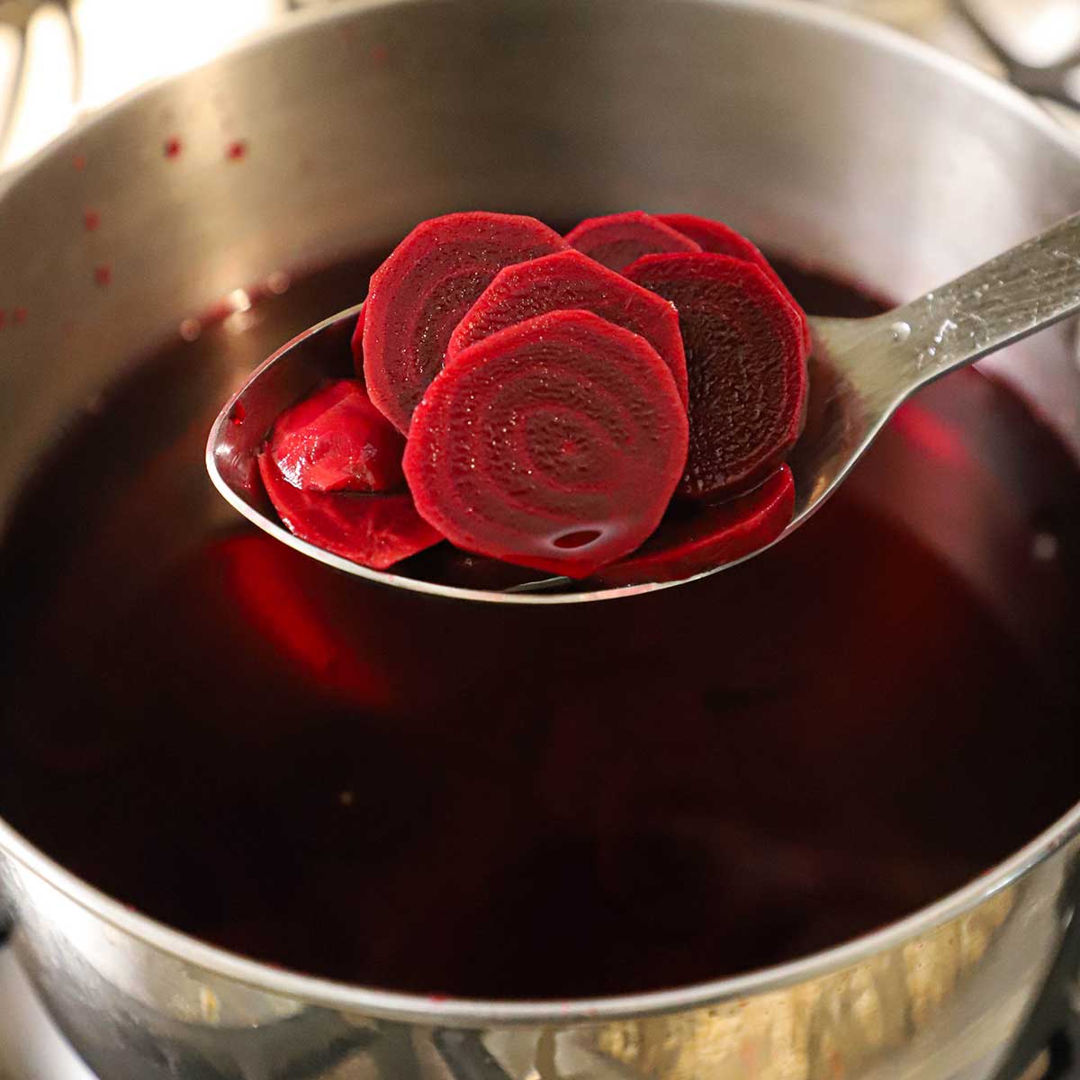 A person holding up a slotted spoon filled with sliced red beets over a pot filled with red beet water.