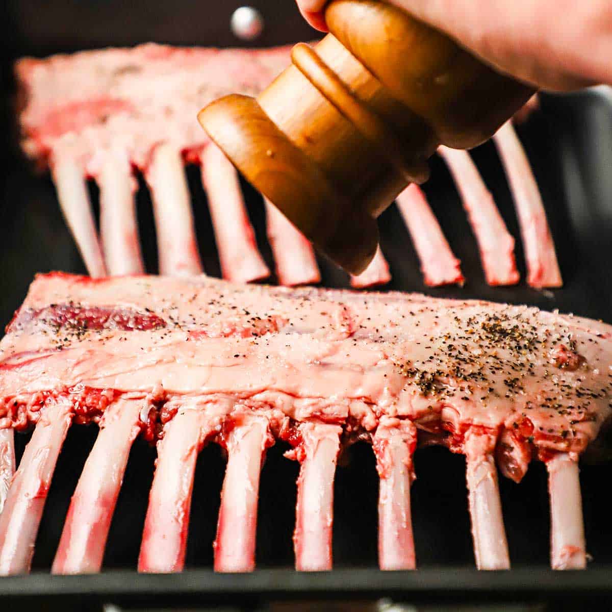 A person using a pepper grinder to season an uncooked rack of lamb in a roasting pan.