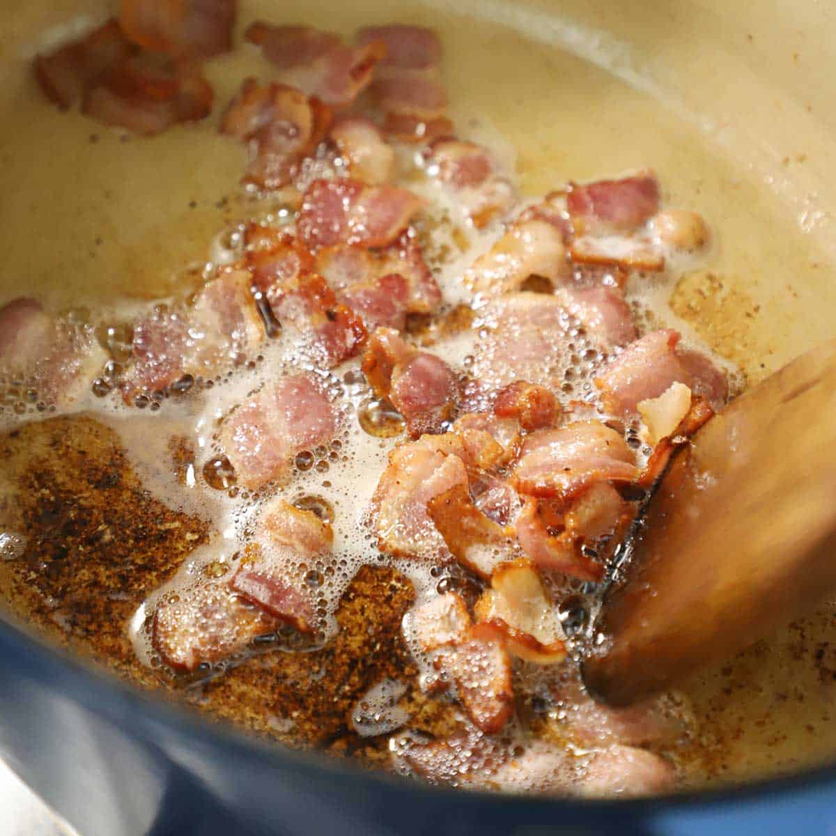Chopped bacon cooking in a large Dutch oven with a wooden spatula stirring it.