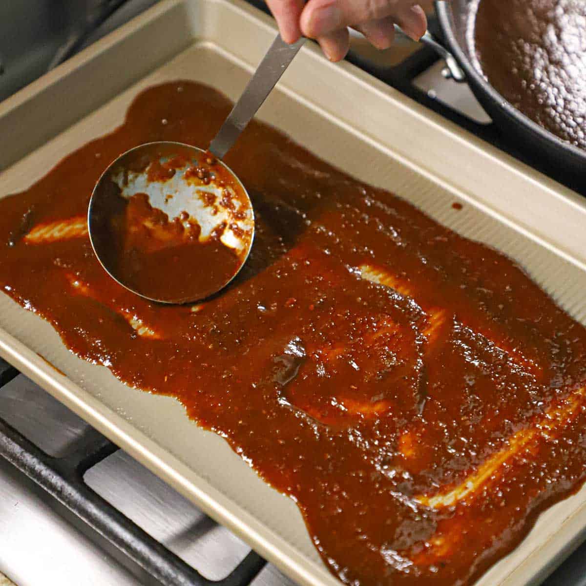 A person using a ladle to spread red enchilada sauce across the bottom of a sheet pan.