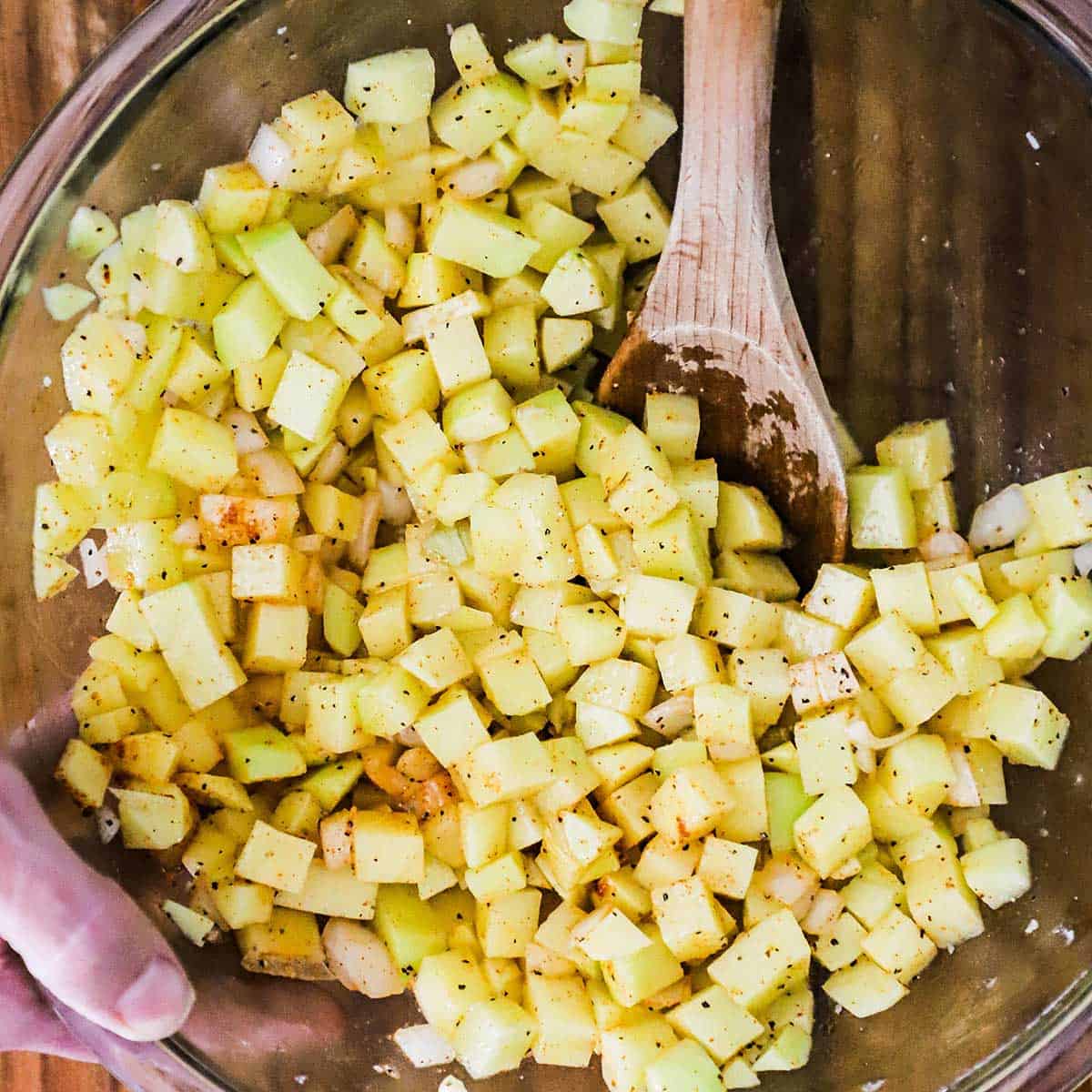Cubed gold potatoes in a large glass bowl being tossed with melted butter, oil, herbs, and seasonings with a wooden spoon.