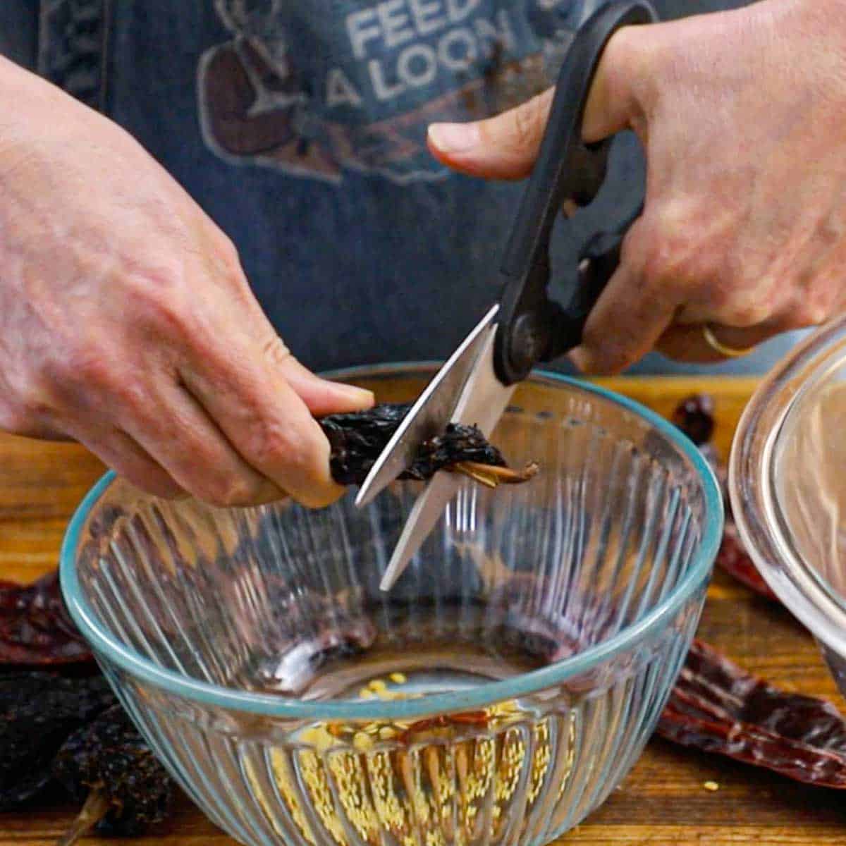 A person using kitchen scissors to cut off the stems from a dried chili over a bowl.