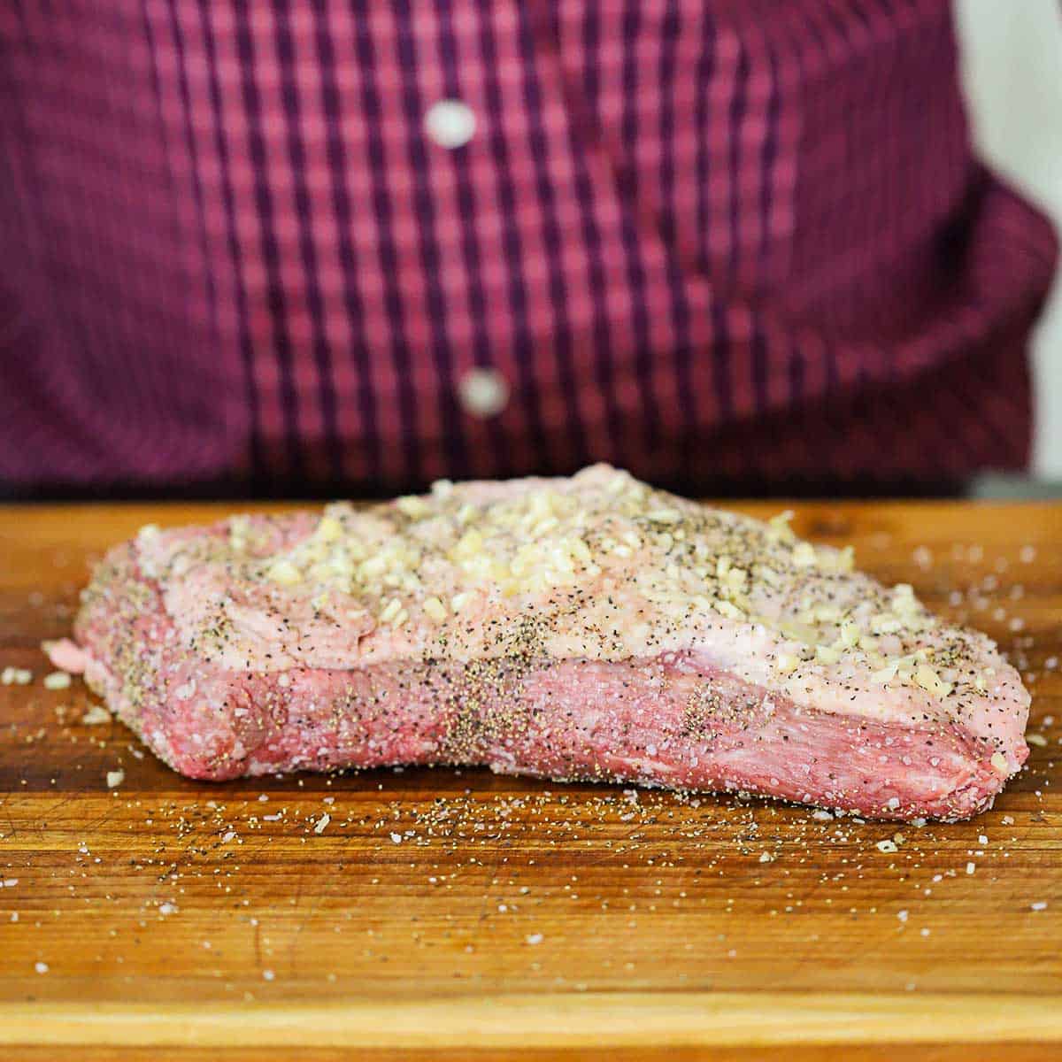 A person standing behind a cutting board with an uncooked seasoned brisket flat on it.