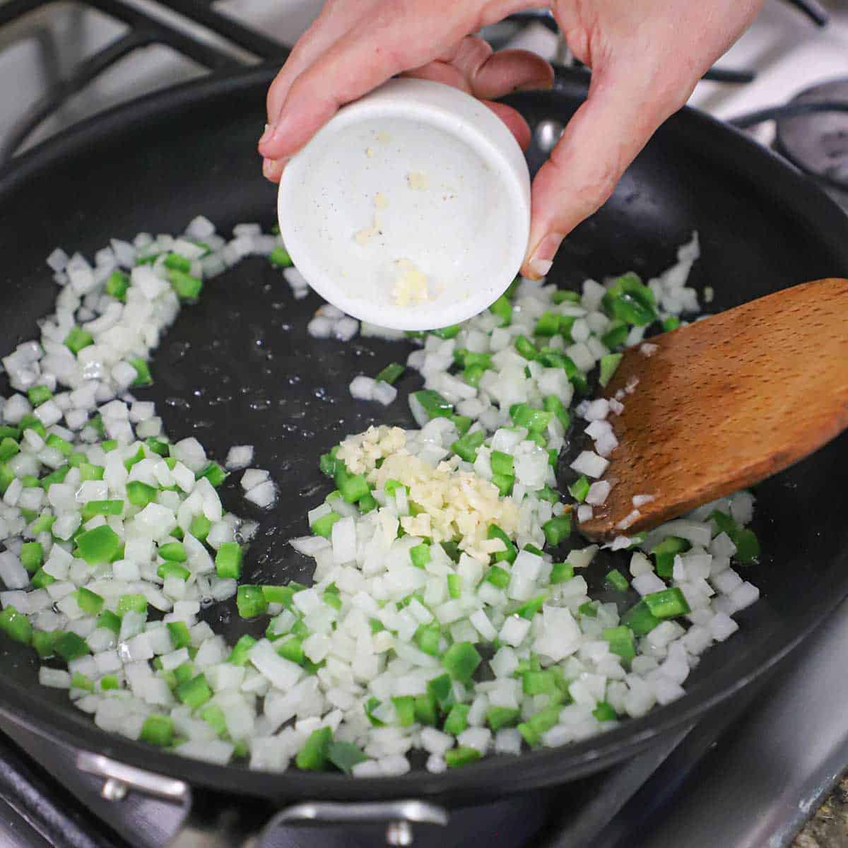 A person dumping minced garlic into a skillet filled with chopped onion and jalapeño being sautéed in olive oil.