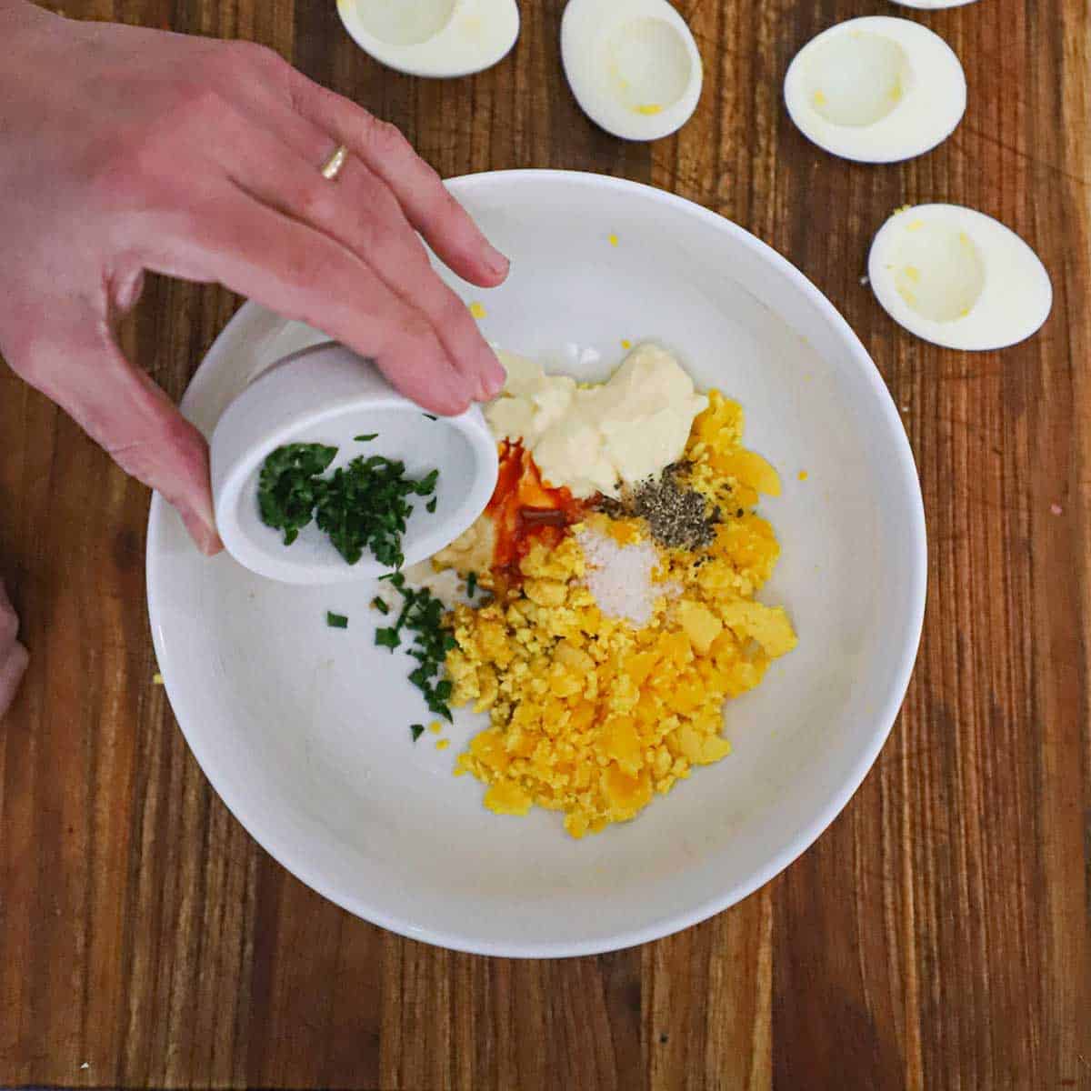 A person transferring chopped chives into a white bowl filled with the ingredients for traditional deviled eggs.