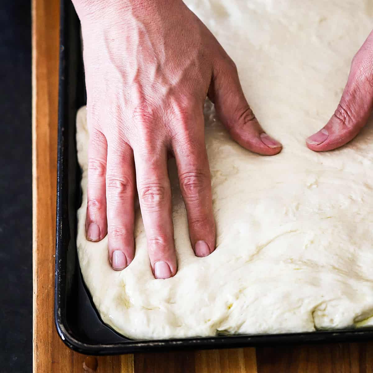 A person user his fingers to stretch focaccia dough into the corners of a baking sheet pan.