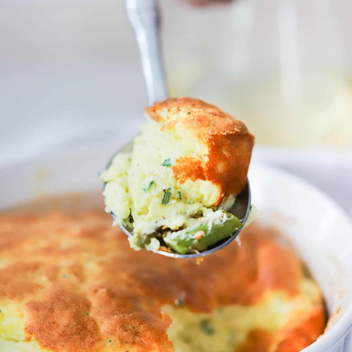 A spoon holding a spoonful of cheese soufflé with asparagus over a baking dish filled with the same.