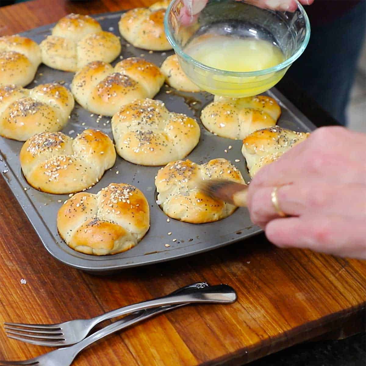 A person brushing melted butter over the tops of freshly baked cloverleaf dinner rolls that are a muffin tin.
