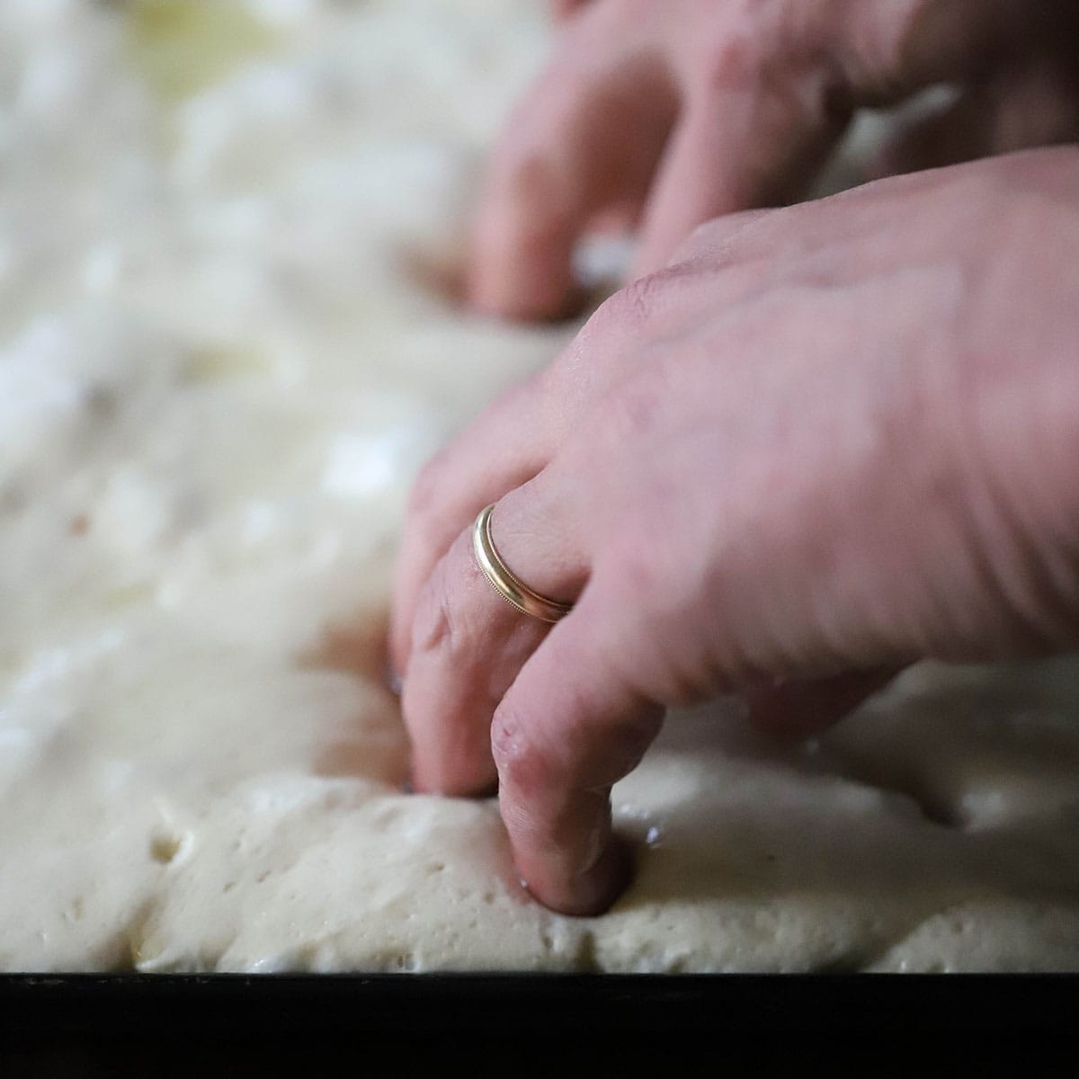 A person pressing his fingers into focaccia dough on a baking sheet causing dimples throughout the uncooked dough.