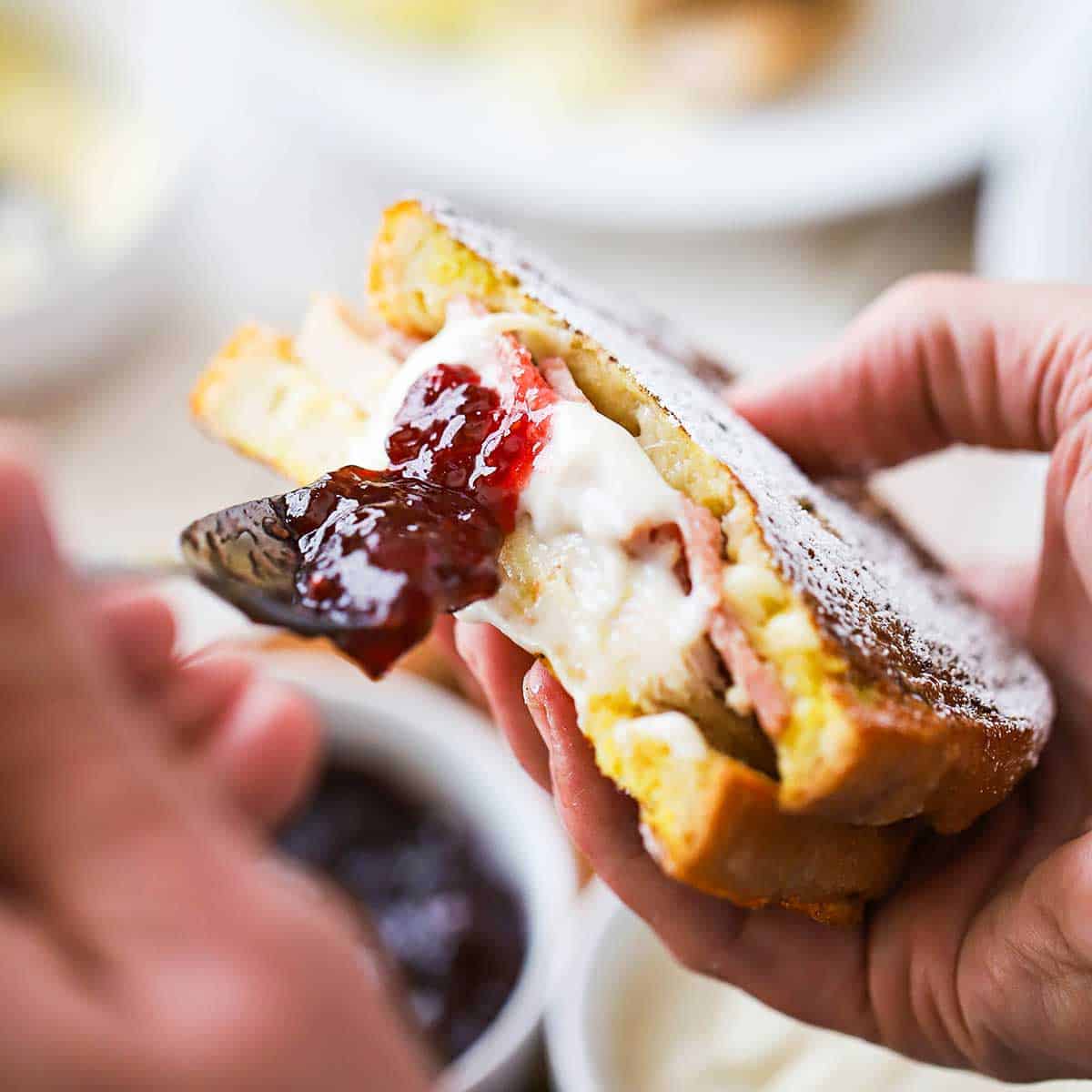 A person smearing fruit preserves onto a Monte Cristo sandwich that has been cut in half.