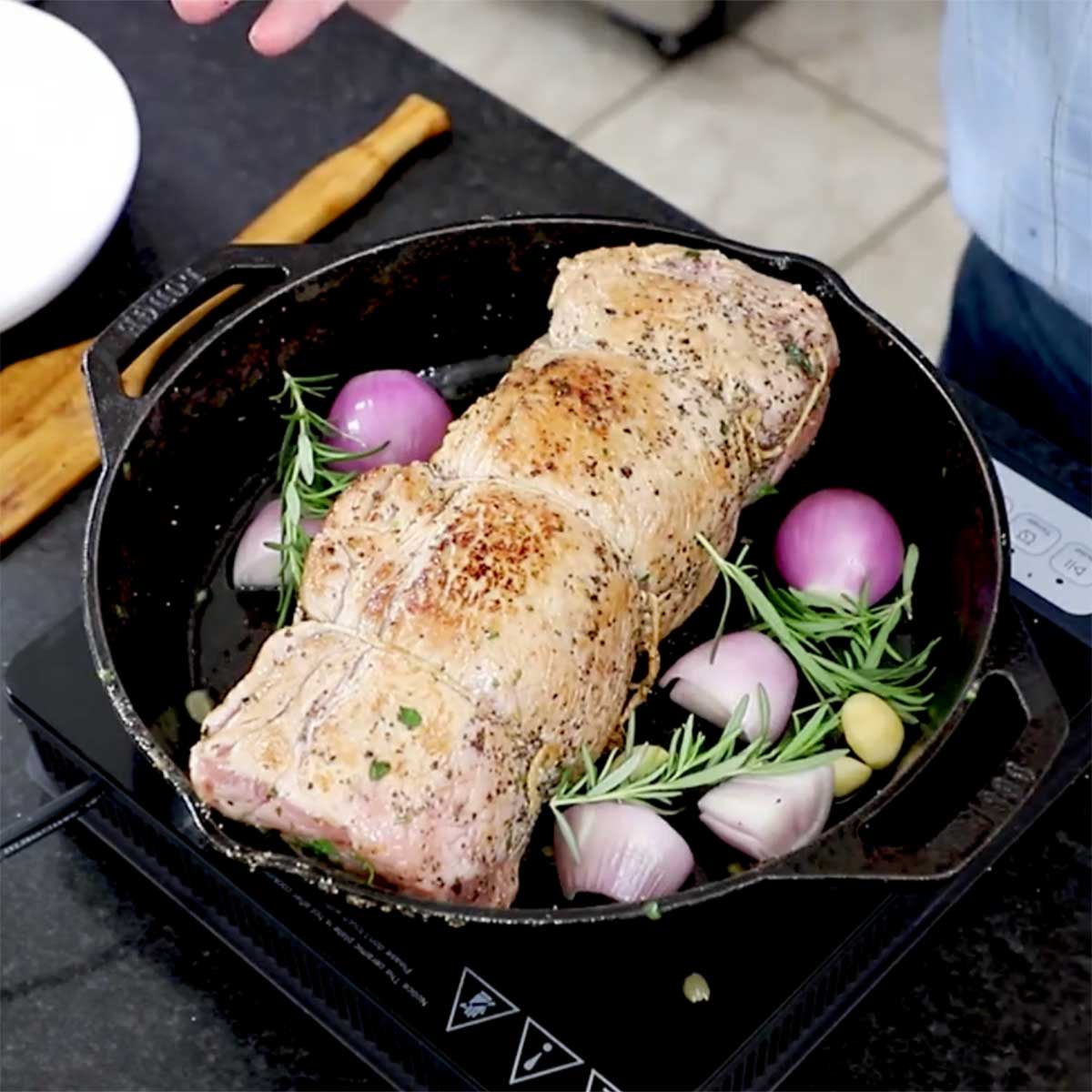 A seared herb-stuffed pork loin resting in a cast-iron skillet with a shallot, garlic, and herbs also in the pan.
