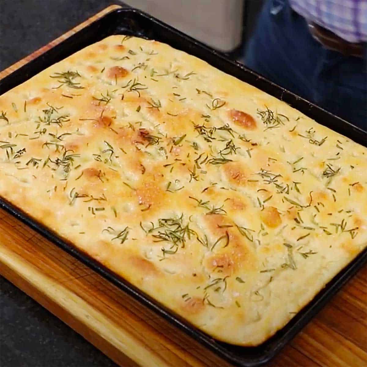A fully baked loaf of focaccia with rosemary and olive oil in baking sheet pan on a cutting board.