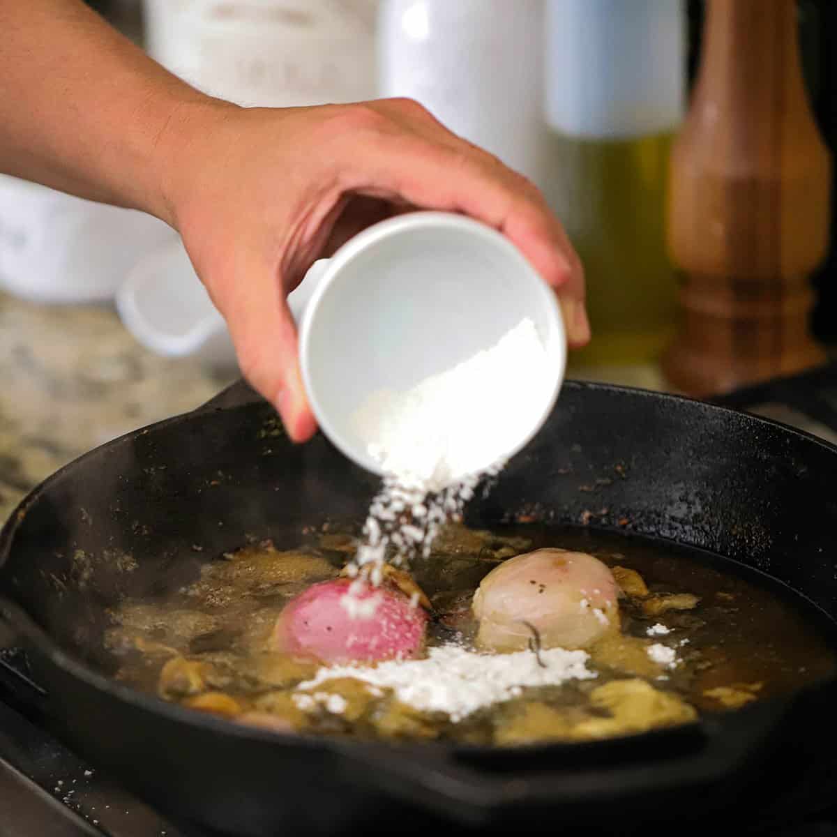 A person adding flour to a skillet that is filled with simmering oil and a shallot and cloves of garlic.
