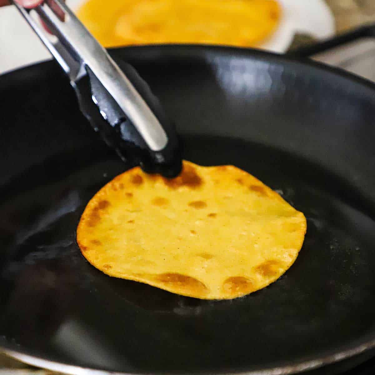 A person using a pair of tongs to lightly fry a corn tortilla in a black skillet filled with a layer of hot canola oil.
