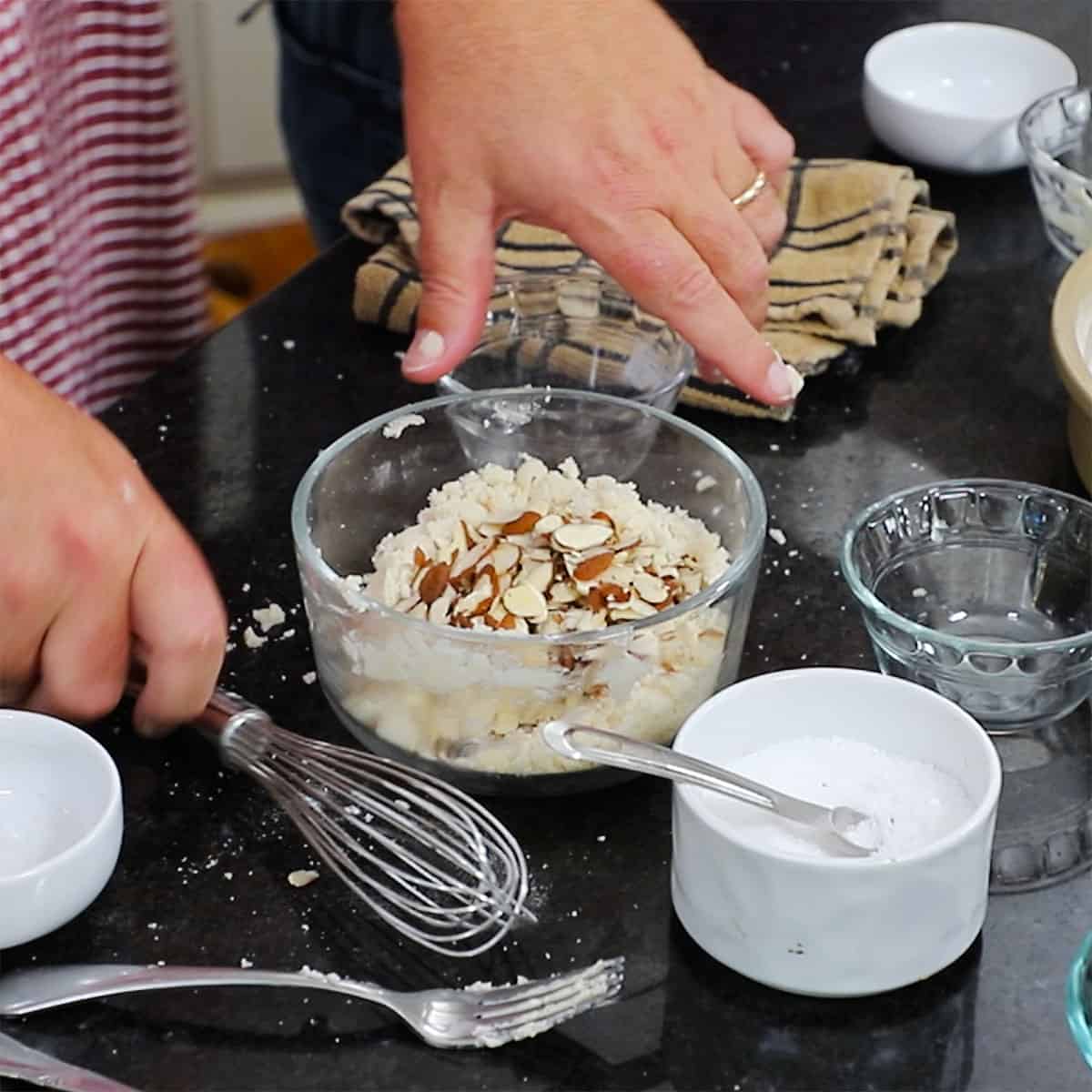 A person adding almond slices into a bowl that has a mixture of softened butter with flour, salt, and sugar in it.