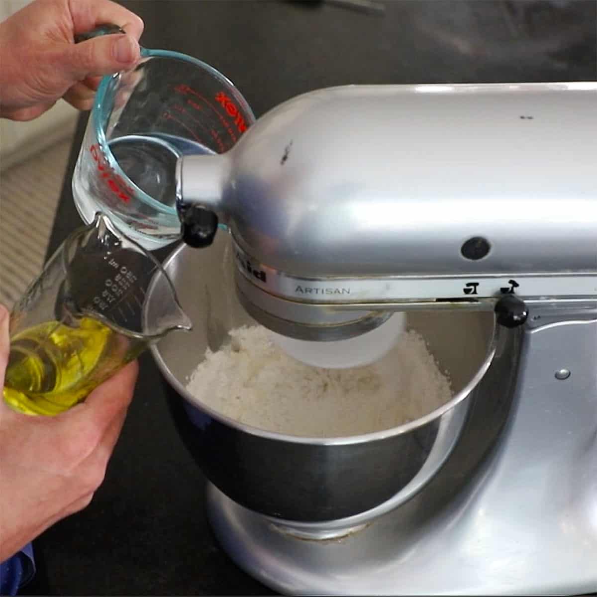 A person pouring hot water from a measuring cup and olive oil from another measuring cup into a mixing bowl filled with flour, yeast, sugar, and salt.