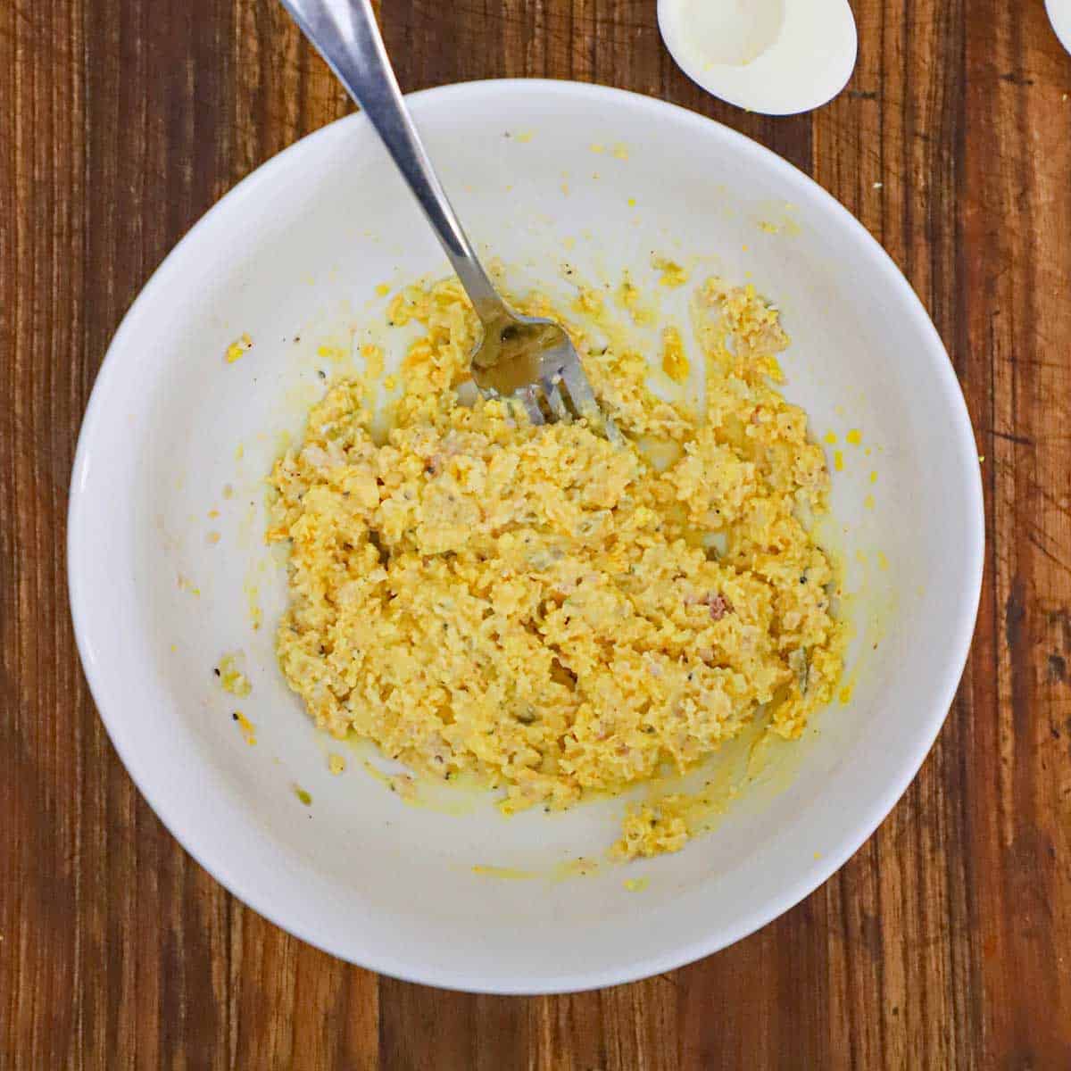 The filling for ham and cheese deviled eggs that have been mashed in a white bowl with the back of a fork.