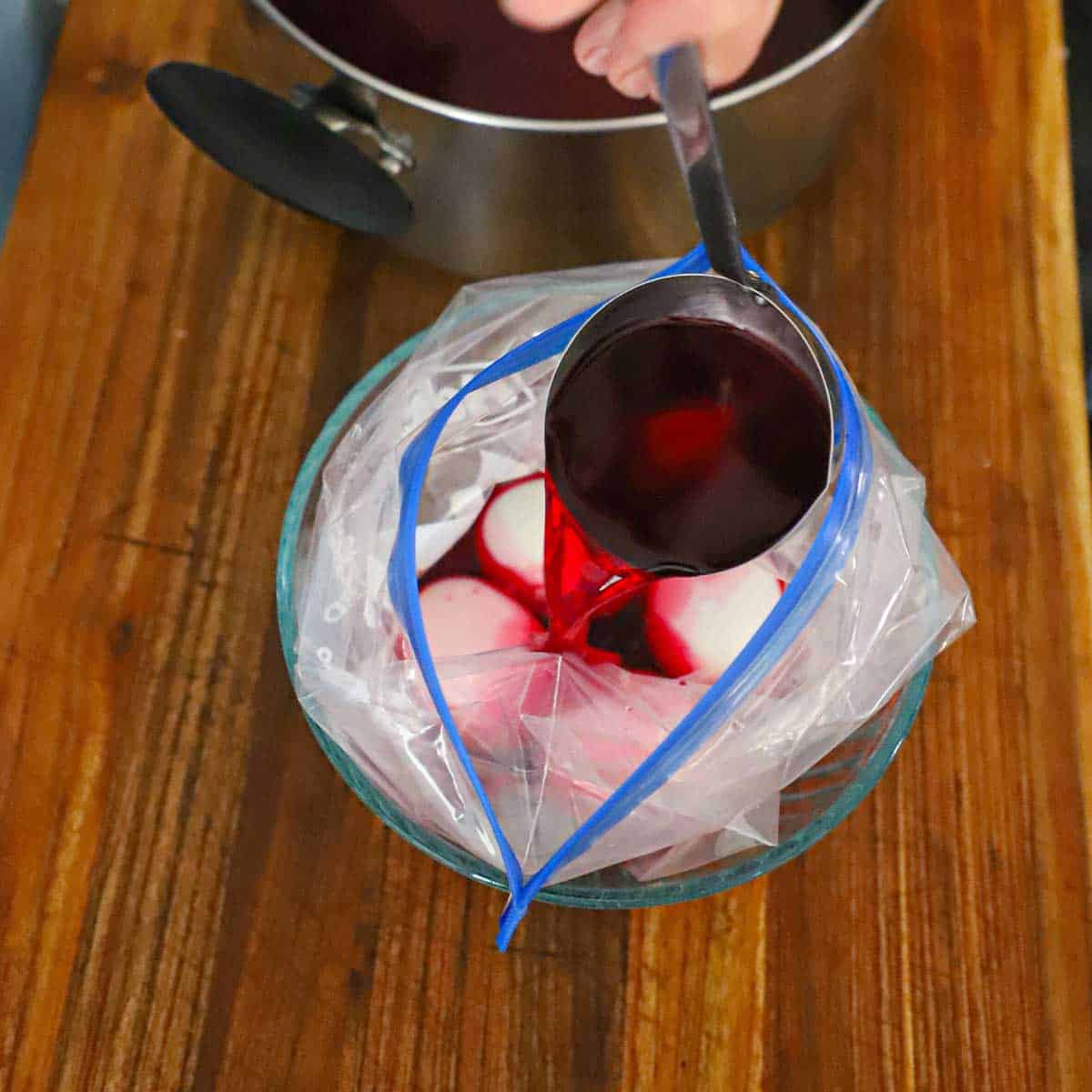 A person ladling red beet water into a plastic baggie that is filled with peeled hard boiled eggs.