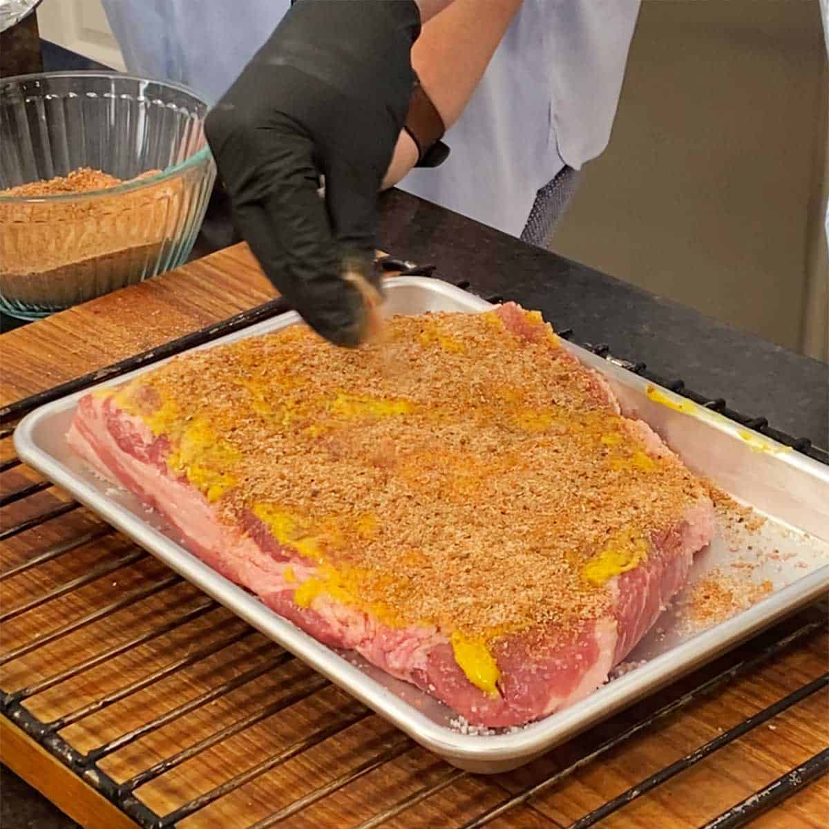 A person with a black latex glove on is sprinkling pork rub all over a pork brisket that has been smeared with yellow mustard.