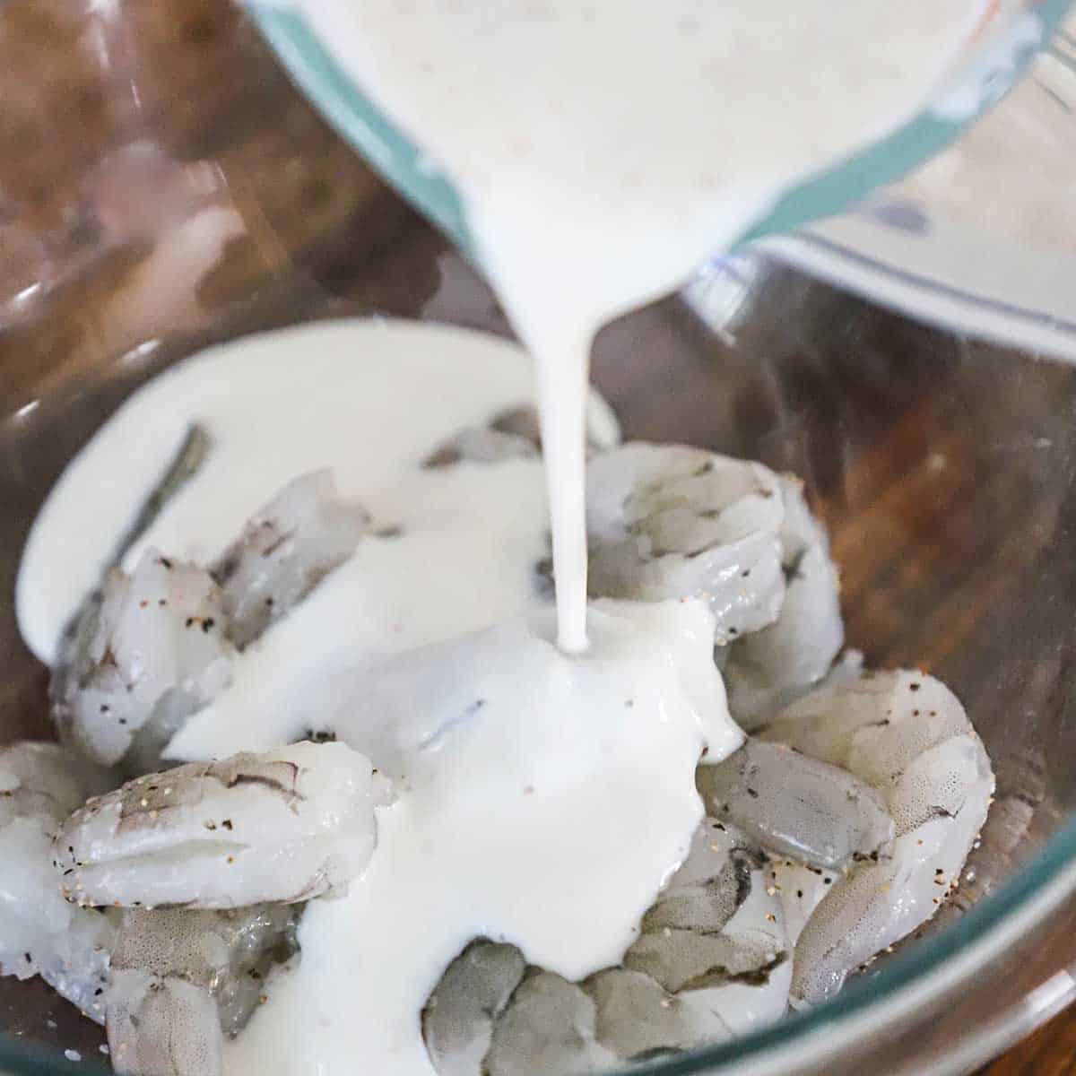 A person pouring buttermilk from a glass measuring cup into a bowl filled with seasoned uncooked shrimp.