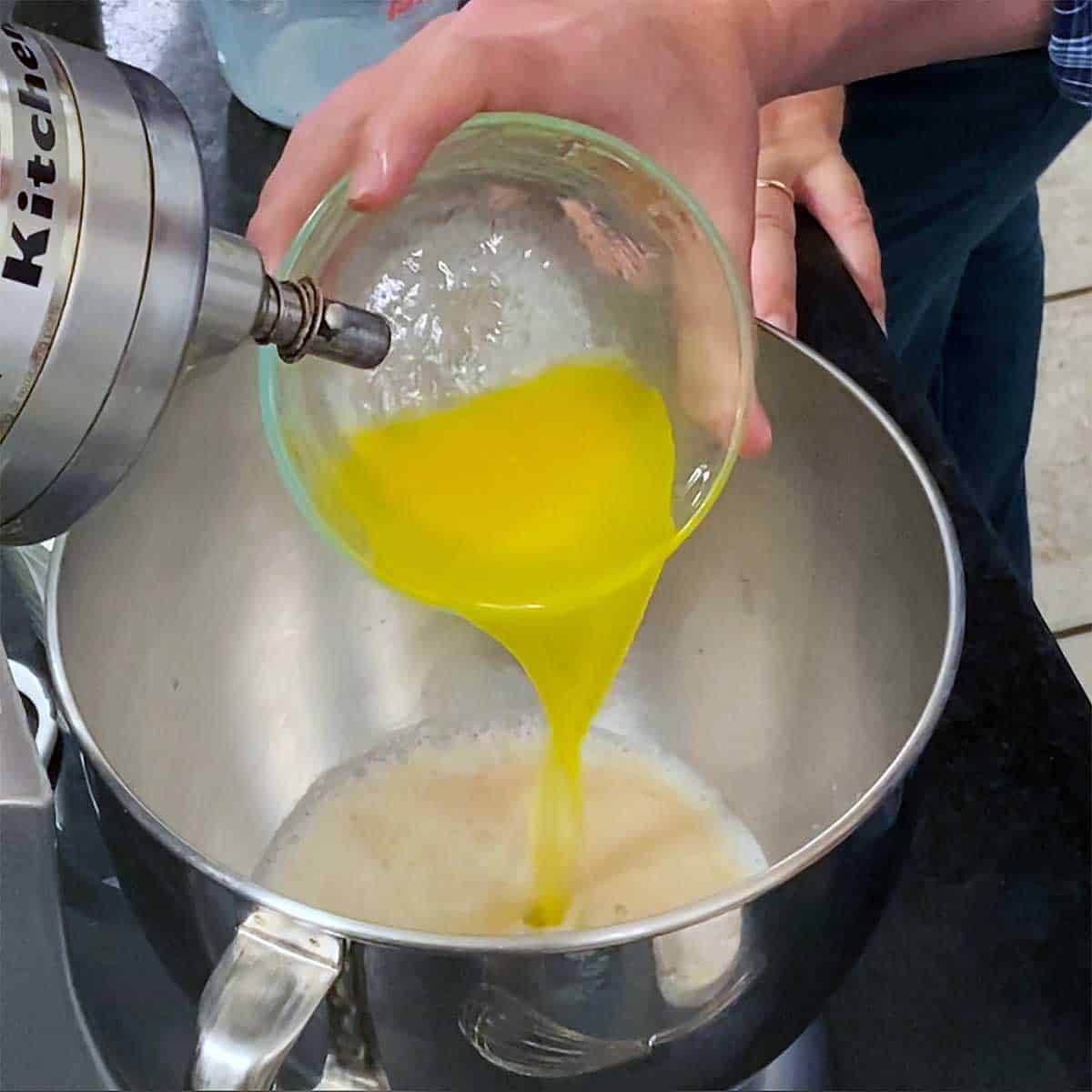 A person pouring melted butter into a bowl of a stand-mixer that is filled with foamy yeast, water, and sugar.