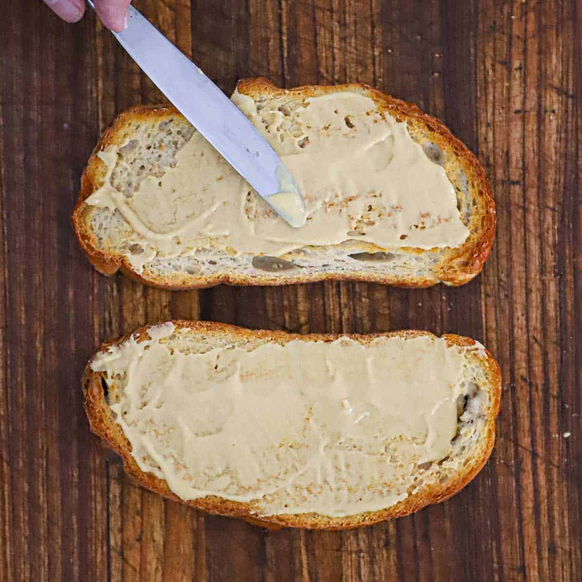 A person using a butter knife to spread Dijon mustard over the tops of two toasted slices of sourdough bread.
