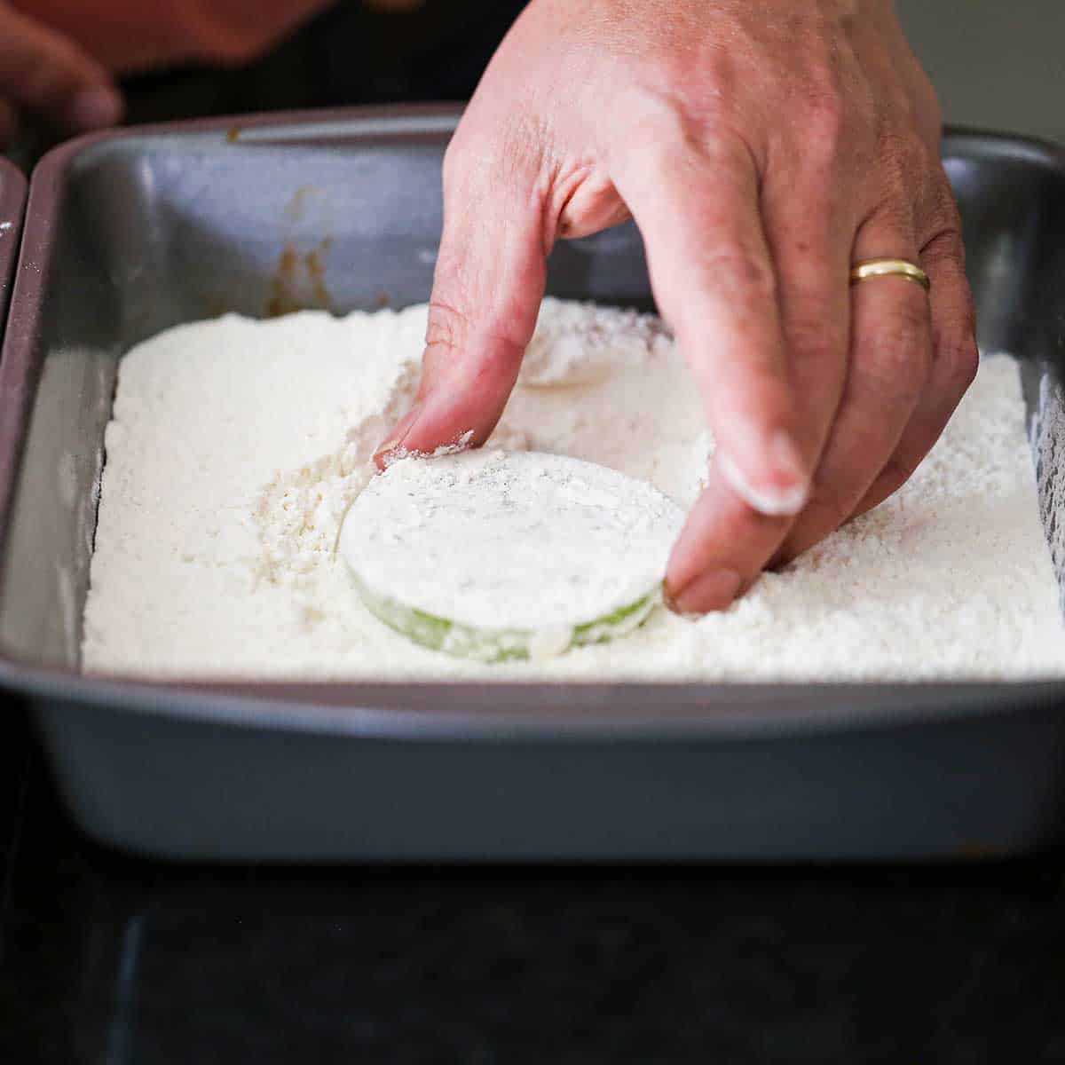 A person dredging a sliced green tomato in all-purpose flour in a square pan.