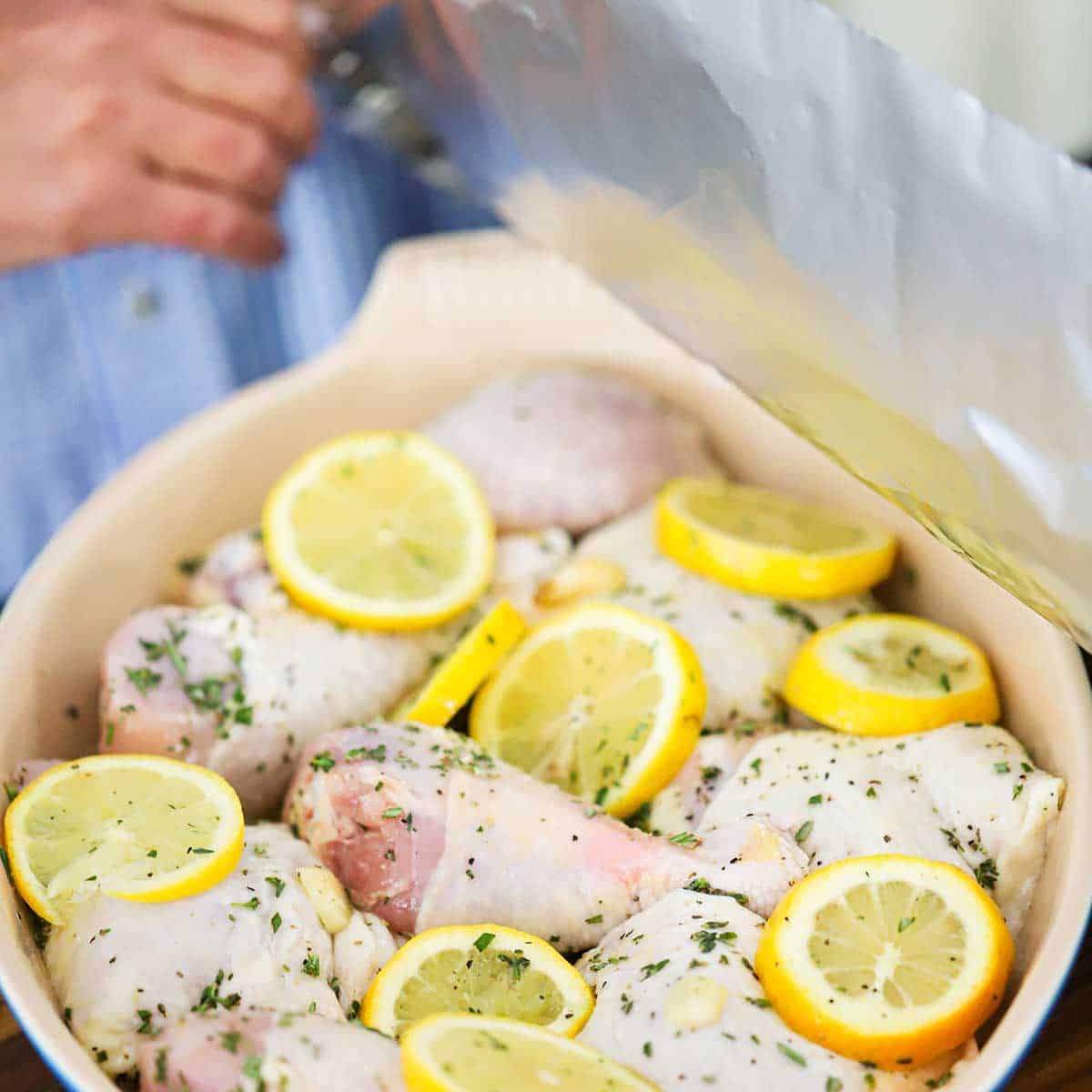 A person placing a large piece of aluminum foil over a baking dish filled with uncooked pieces of chicken, lemon slices, and herbs.