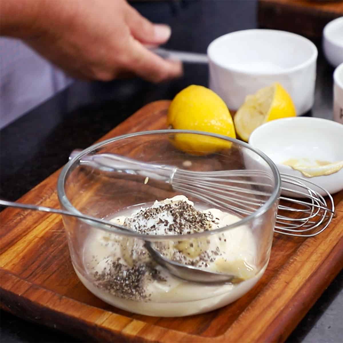 A glass bowl filled with mayonnaise, Dijon mustard, salt, and pepper with halved lemons next to it on a cutting board.
