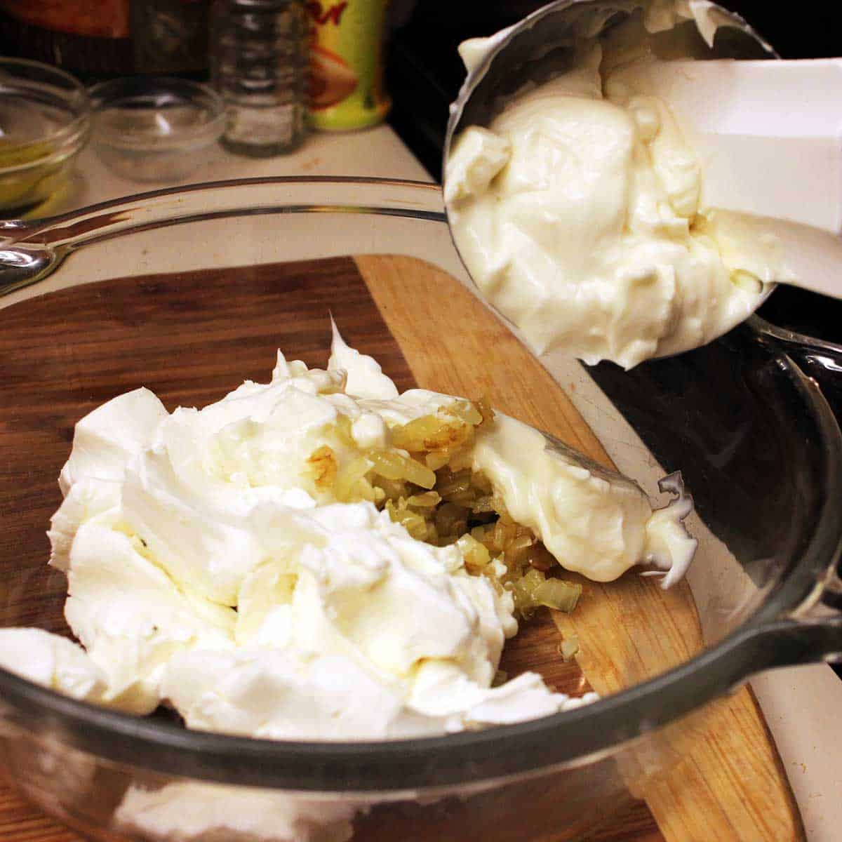 A person adding a cup of mayonnaise into a bowl filled with softened cream cheese and sautéed onions.