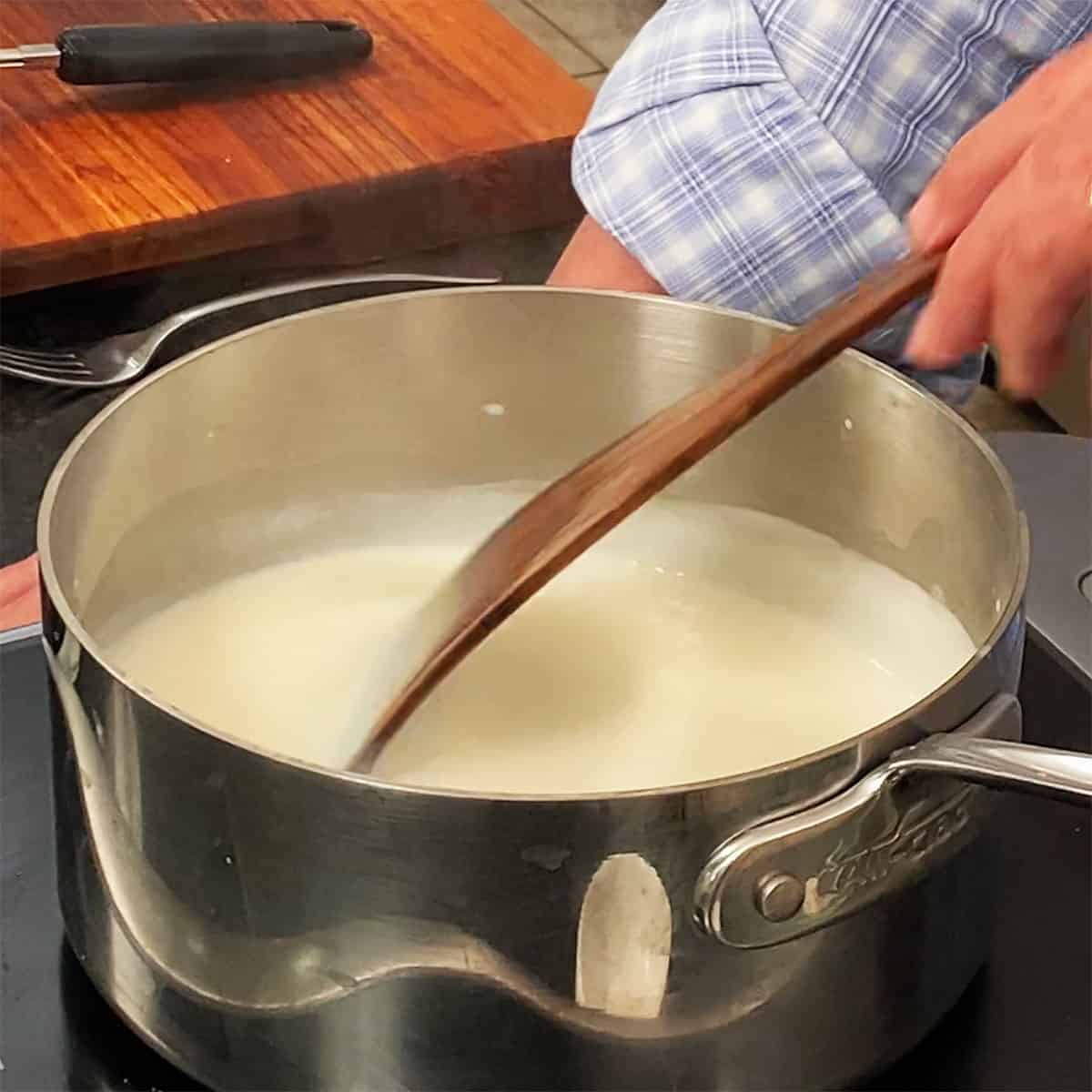 A person using a wooden spatula to stir a vanilla custard sauce in a saucepan.