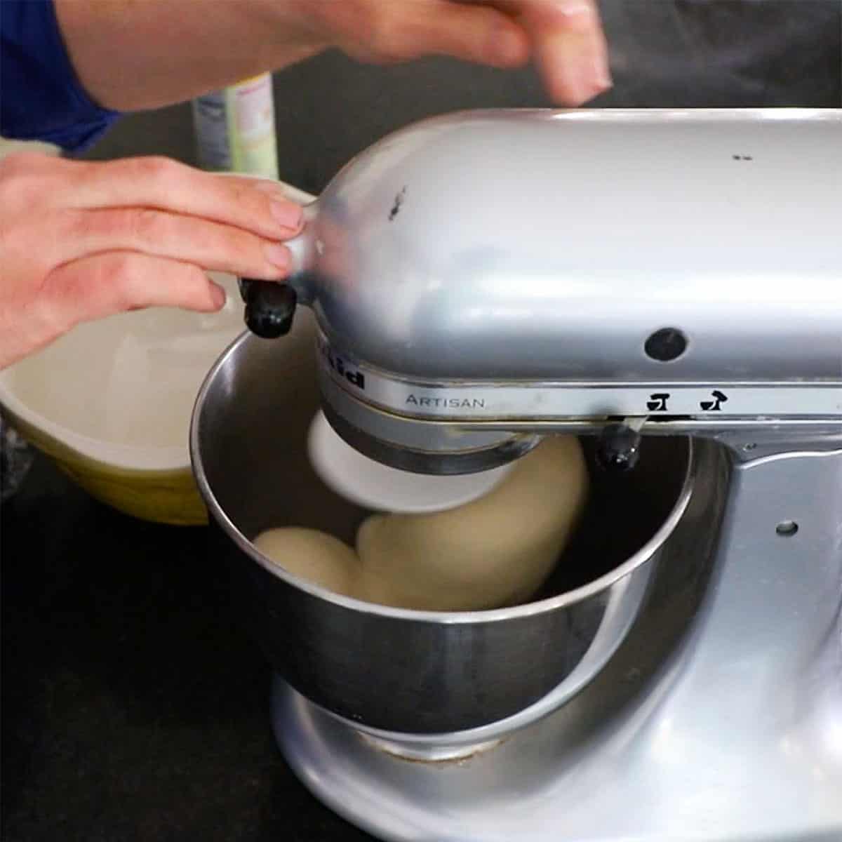 A person standing in front of a stand-mixer that is being used to knead dinner roll dough in the bowl until smooth.