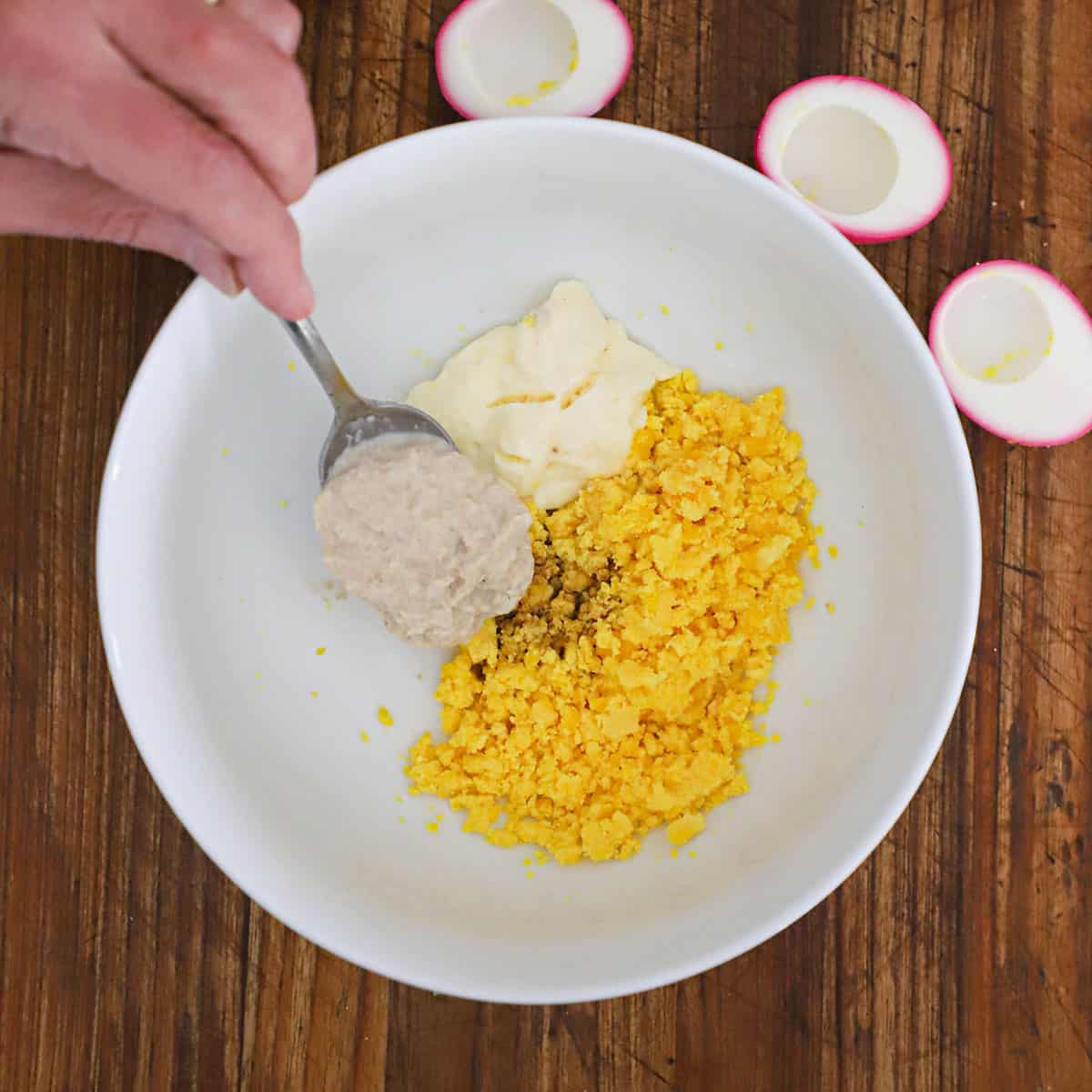 A person adding a spoonful of horseradish sauce into a bowl filled with mashed egg yolks and mayonnaise.