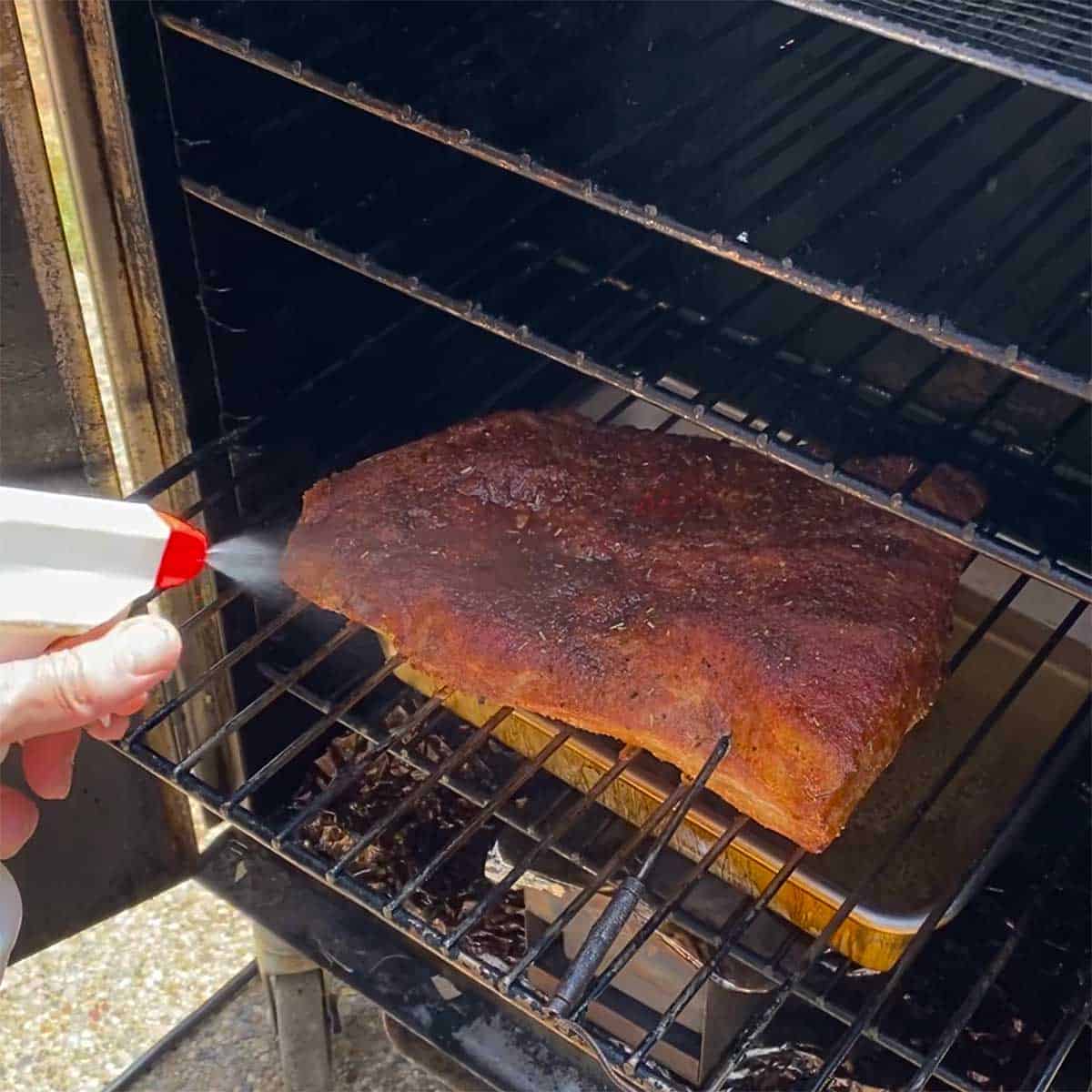 A person using a squirt bottle to spray apple cider vinegar all over a pork brisket that is being smoked in an electric smoker.