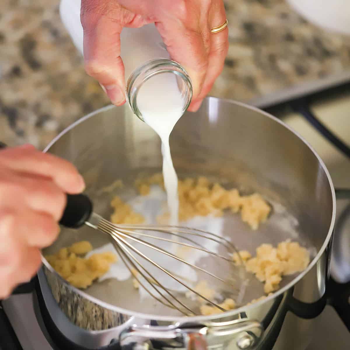 A person pouring milk from a small milk bottle into a silver saucepan with a roux in the bottom of it.