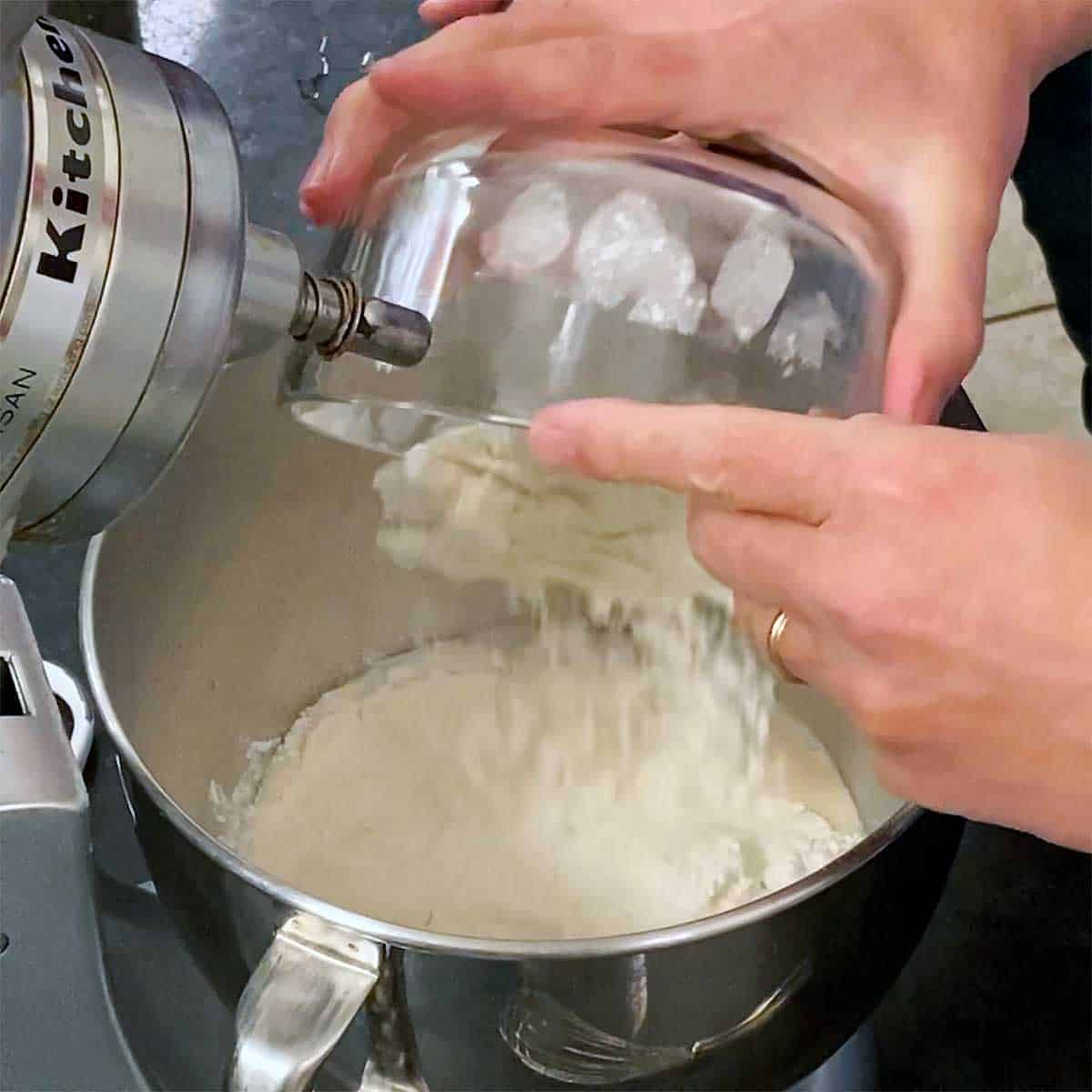 A person dumping all-purpose flour from a glass bowl into a mixer filled with a wet yeast mixture.