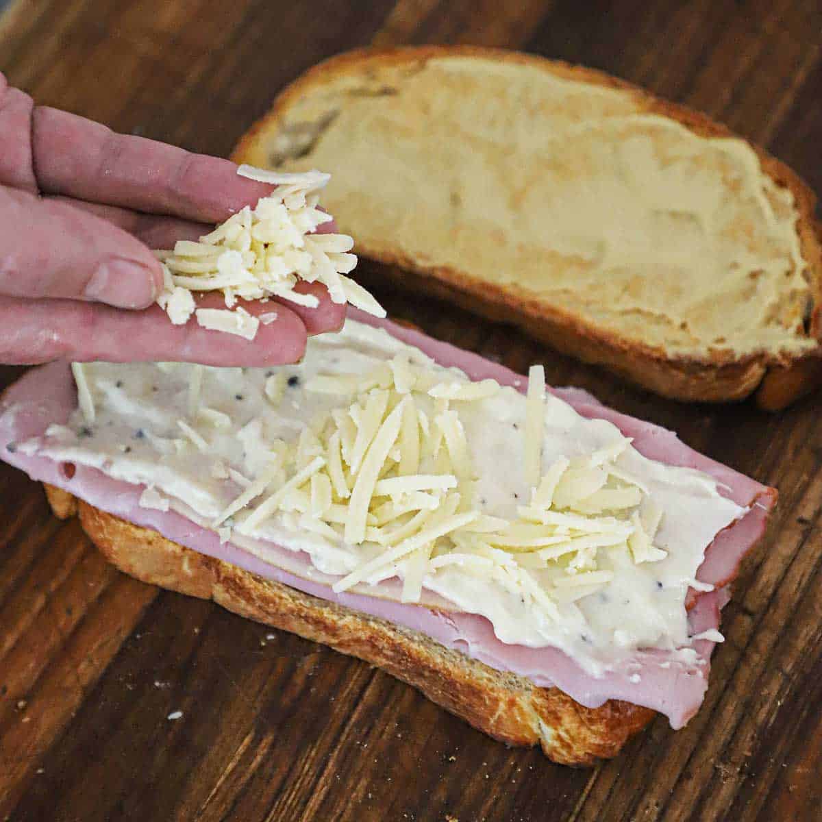 A person sprinkling grated Gruyere cheese onto the bottom half of a Croque Madame sandwich that included bread, sliced ham, and mornay sauce.