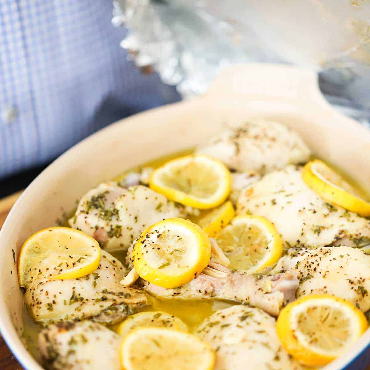 A person removing a large piece of aluminum foil from a baking dish revealing chicken pieces that have been roasted with lemon slices, herbs, oil, and white wine.