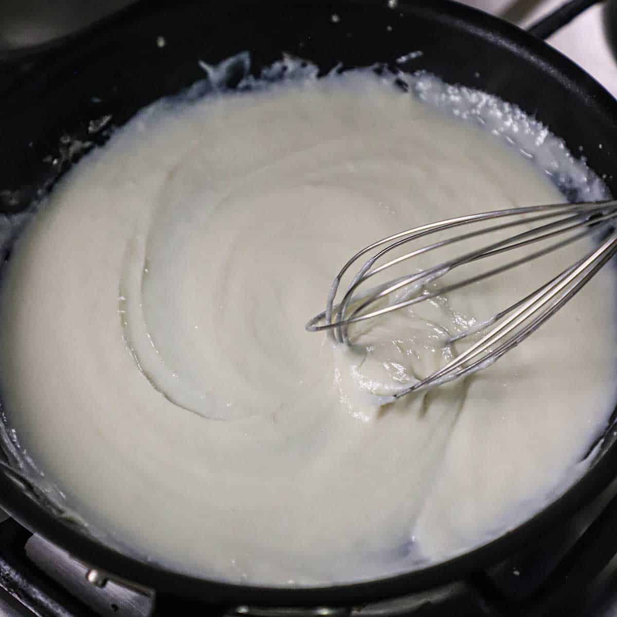 A person whisking a béchamel sauce in a large black skillet.