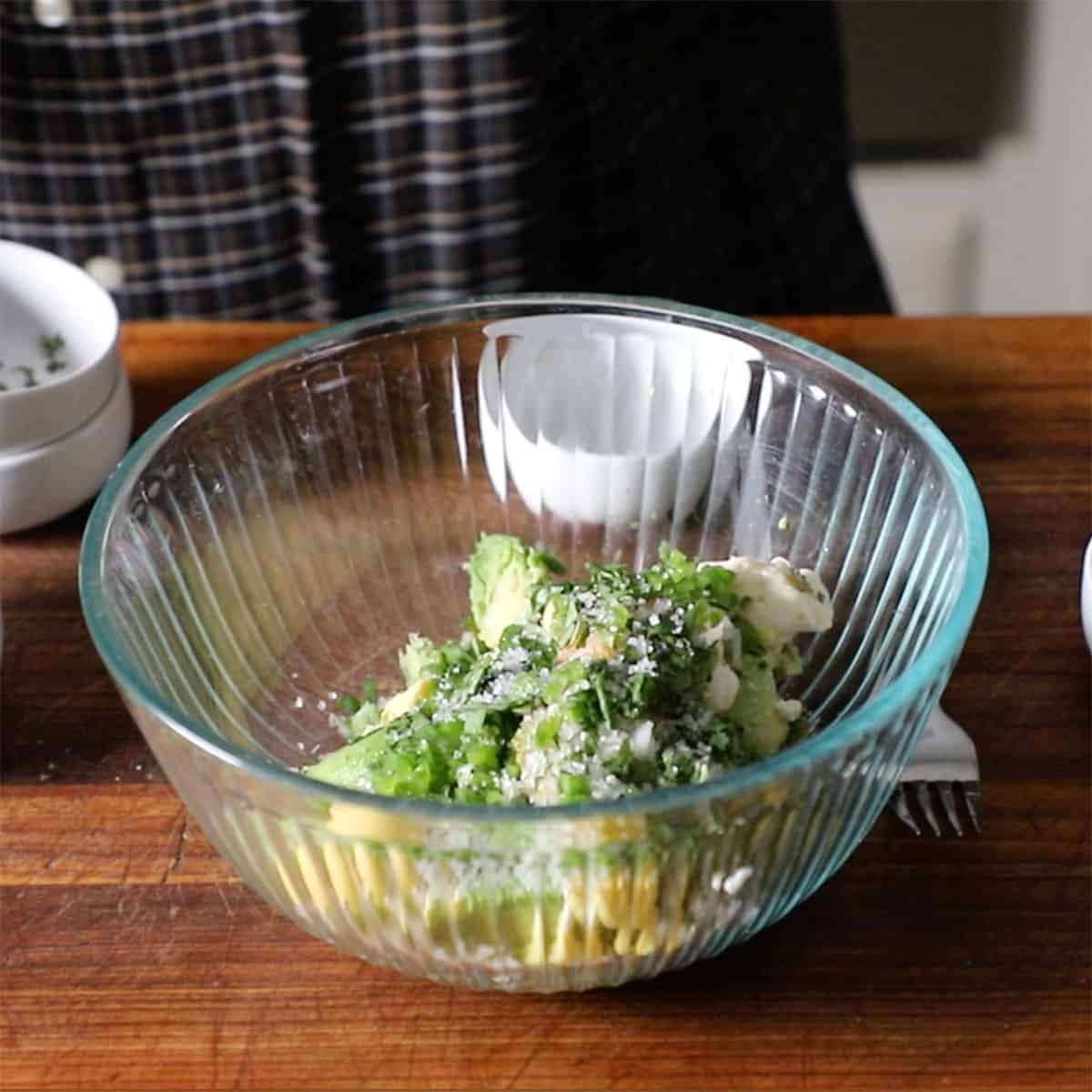A glass bowl filled with hard boiled eggs yolks, avocado chunks, and herbs all sitting on a wooden cutting board.