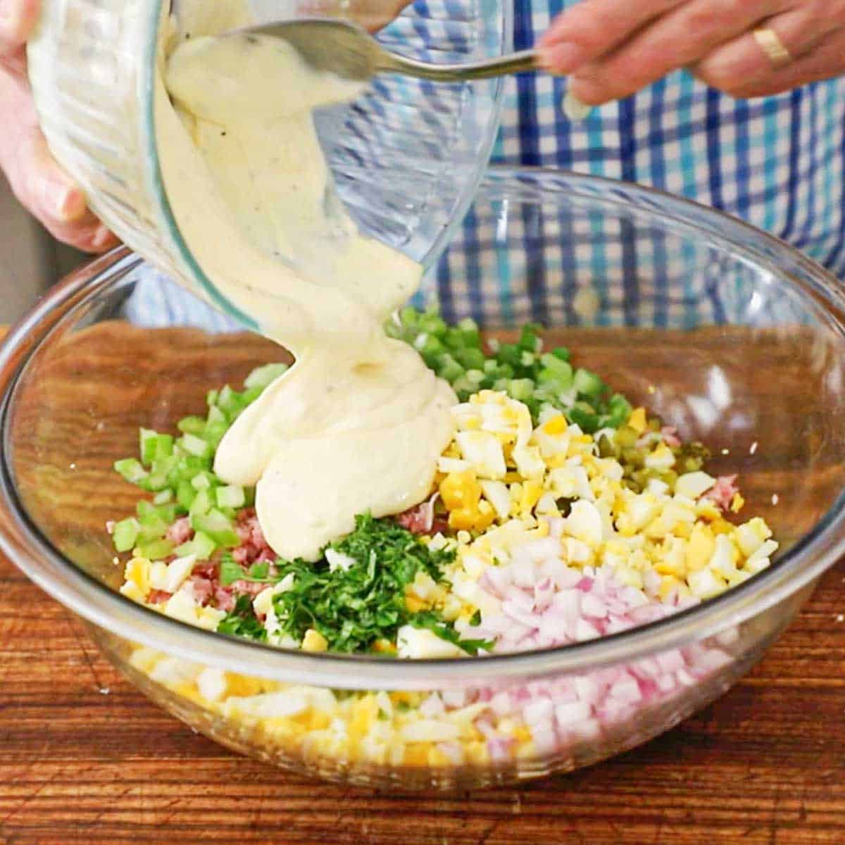 A person pouring a mayonnaise and Dijon dressing from a small glass bowl into a large bowl filled with the ingredients for ham salad.