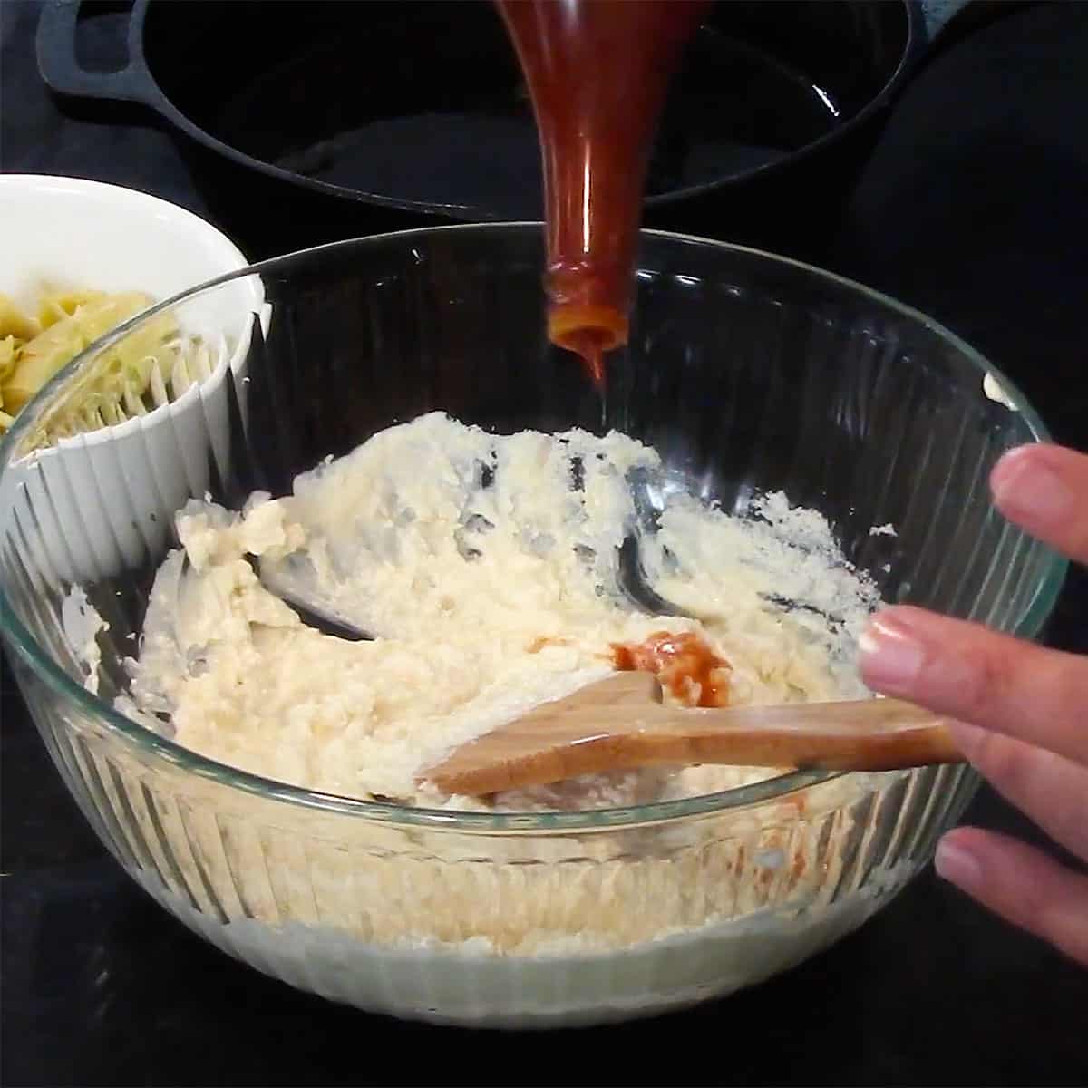 A person adding splashes of hot sauce from a bottle into a glass bowl filled with a mixture of cream cheese, mayonnaise, and sautéed onions.