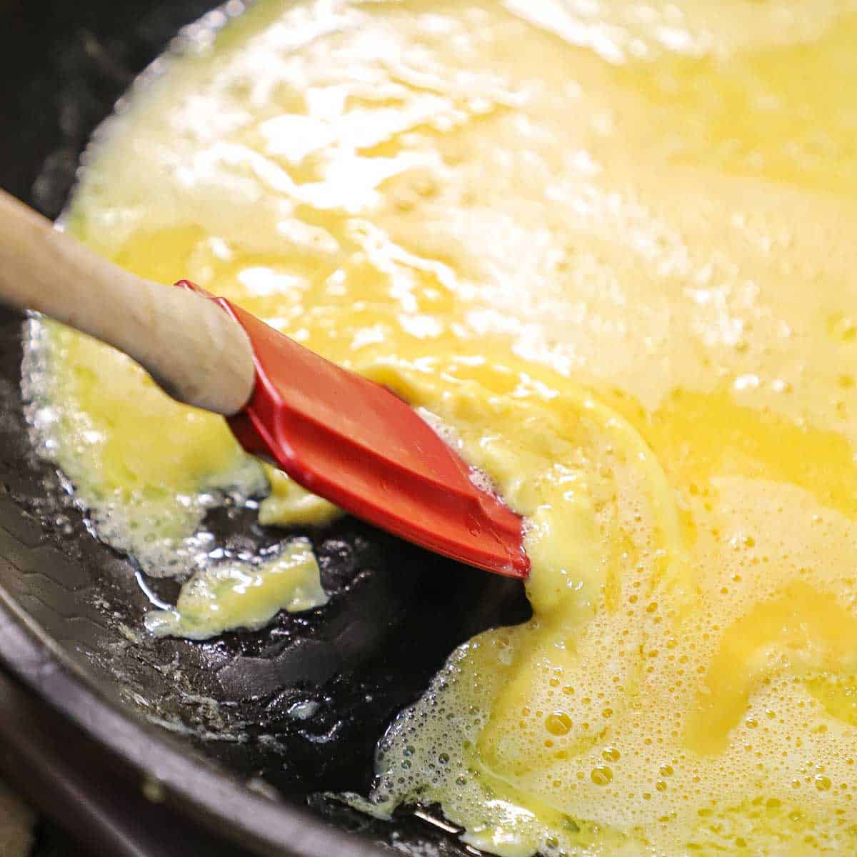 A person using a red spatula to gently move slightly set eggs from the outer edges in a large non-stick skillet.
