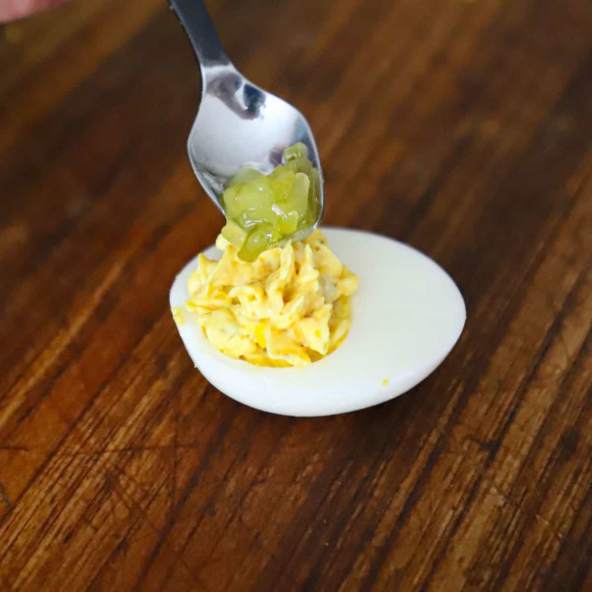 A person using a small spoon to place pickle relish on top of a ham and cheese deviled egg.